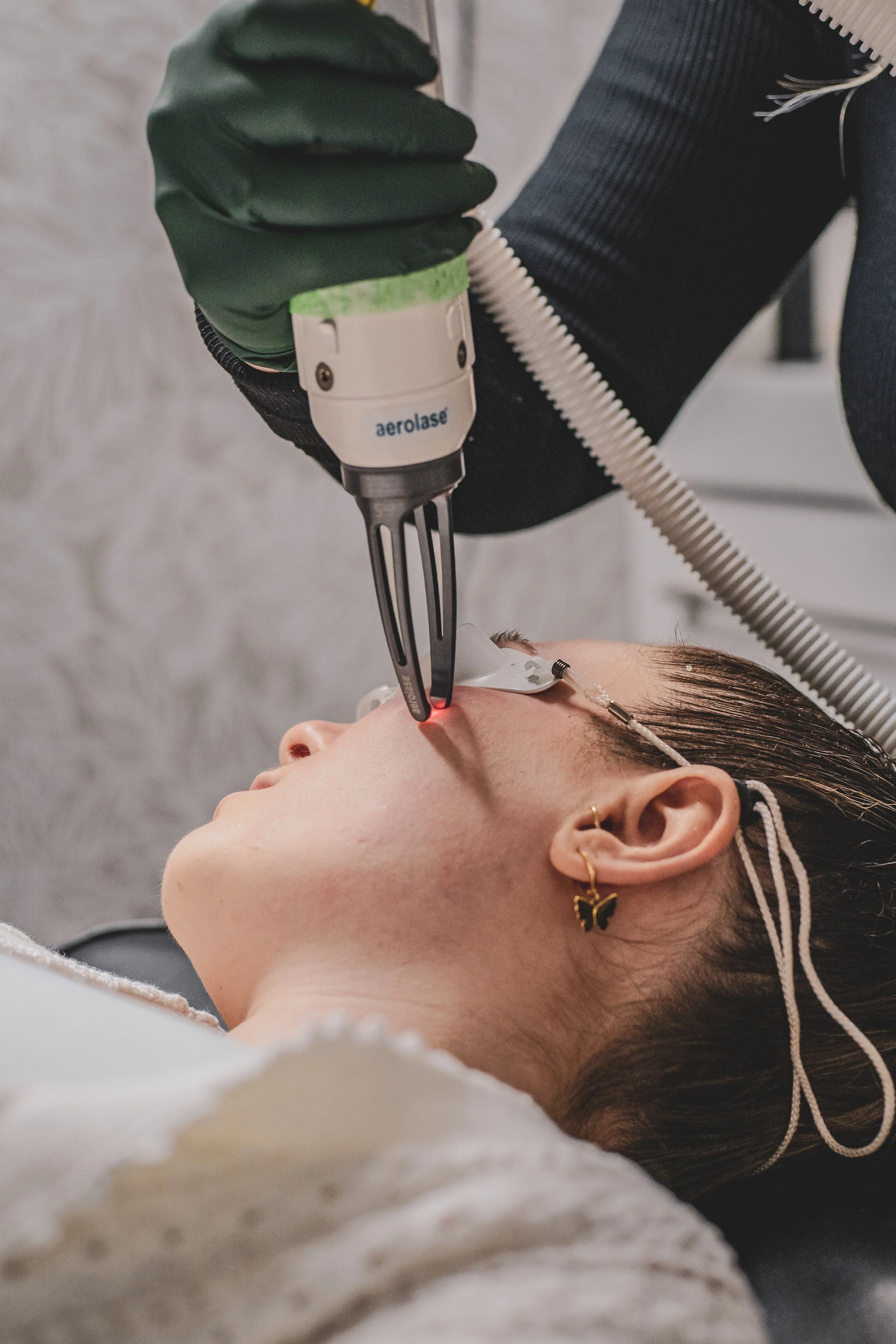 A person receiving a laser treatment for skin concerns. The aerolase laser is held next to their face while they lay on a bed in a salon.