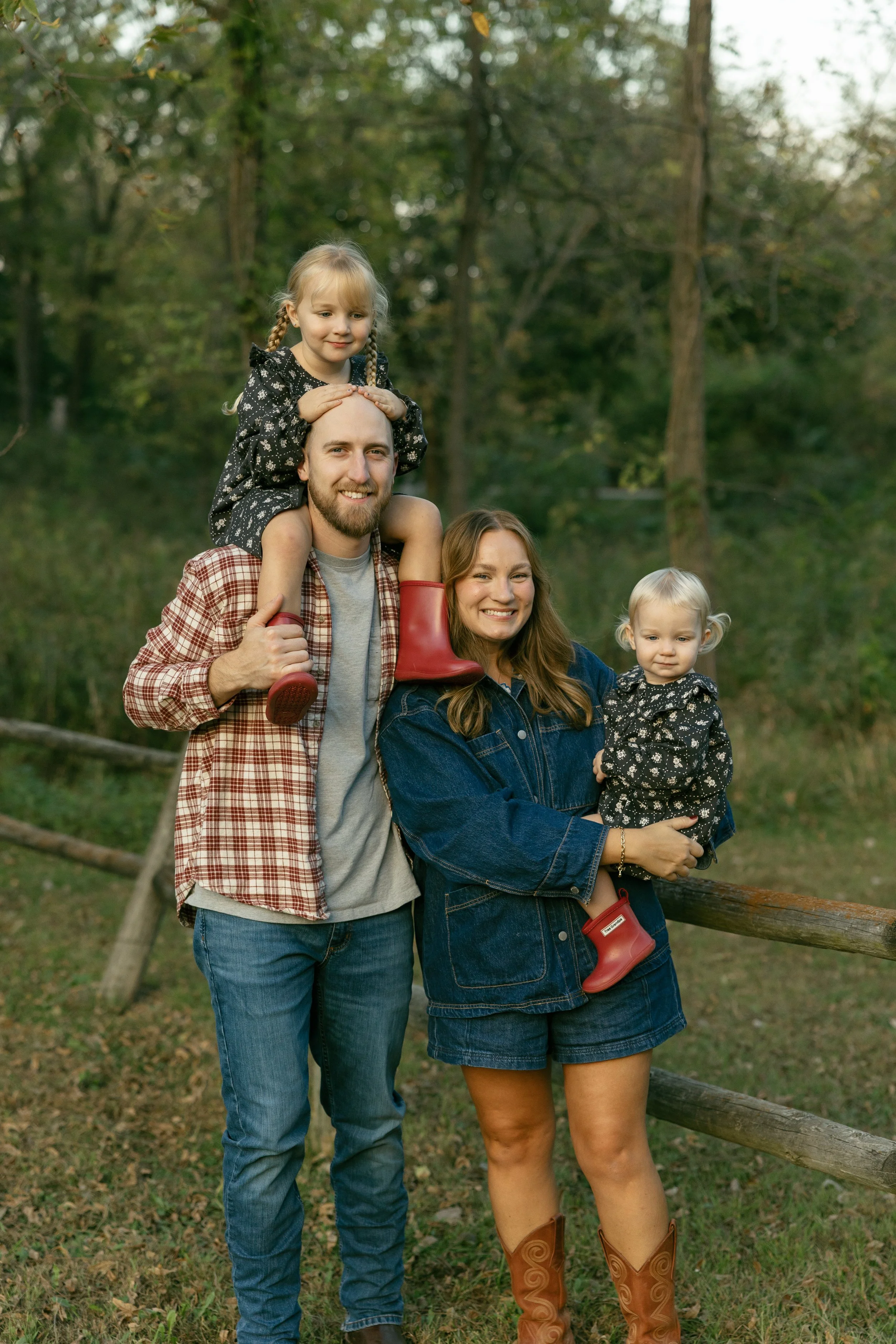 Family of four posing outdoors in a wooded area. The father carries a young girl on his shoulders, and the mother holds a toddler girl. All are smiling and casual dressed.