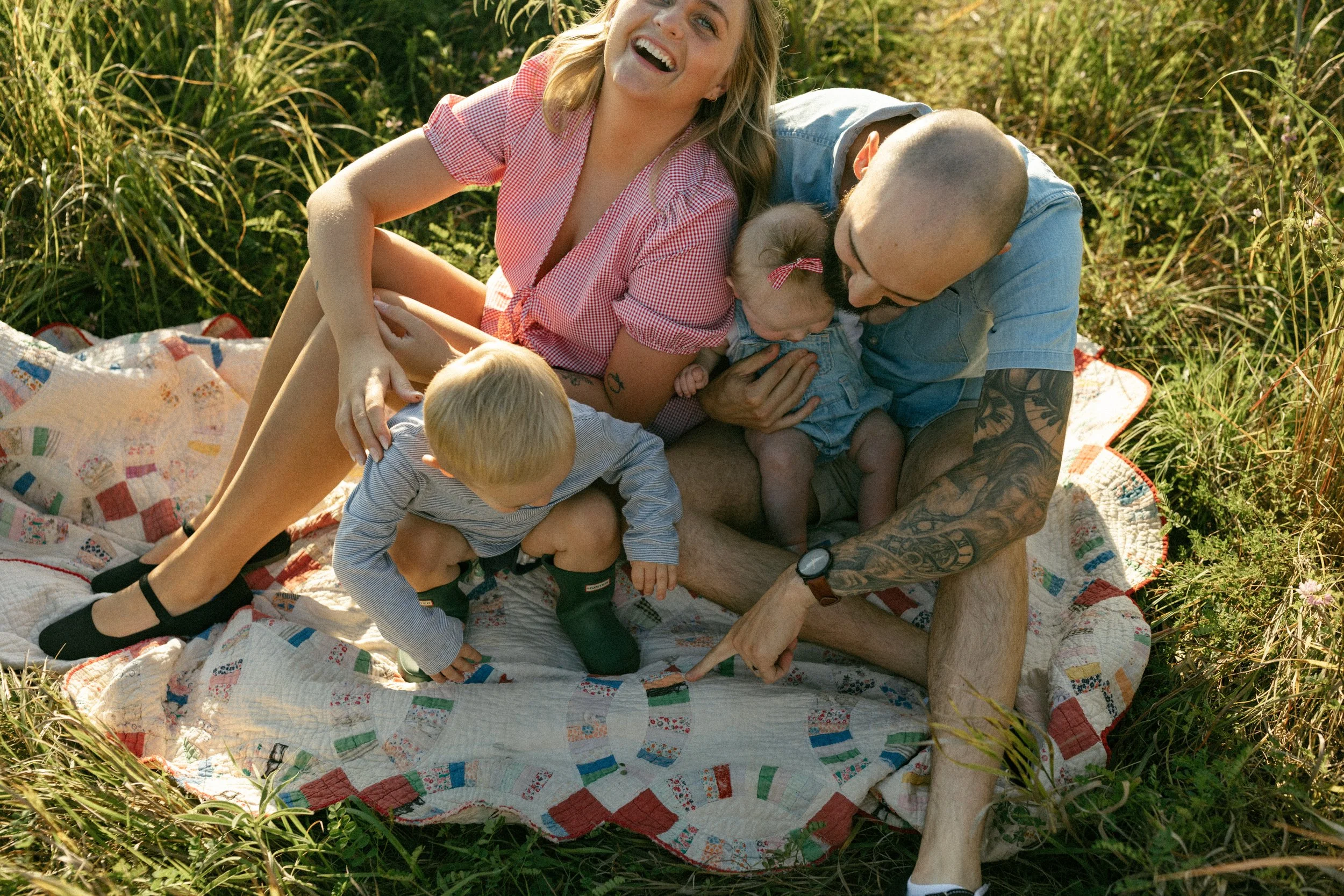 A family of four with two young children sitting on a quilt outdoors, surrounded by tall grass and plants, enjoying a sunny day. The mother and father are interacting with the children, who are climbing on their laps.