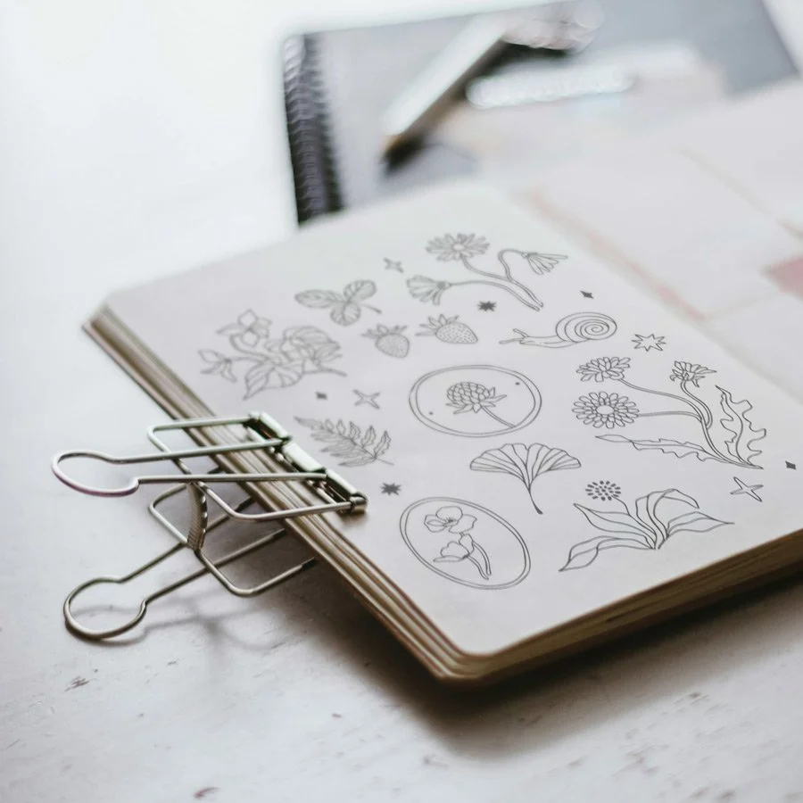 A sketchbook with botanical and insect sketches open on a table, with a binder clip attached to the corner.