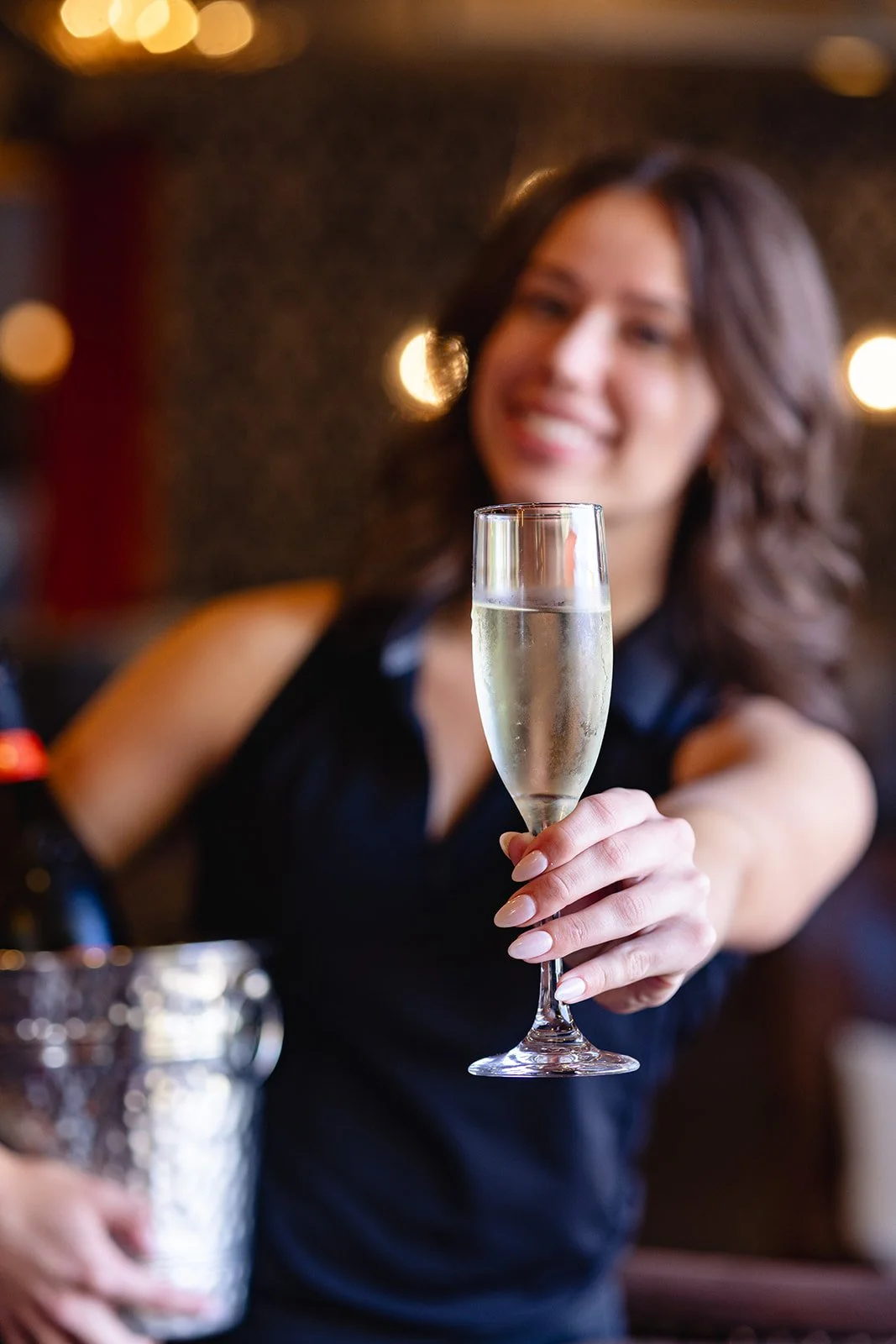 Girl in black dress smiling while holding a glass of champagne in a dimly lit setting.