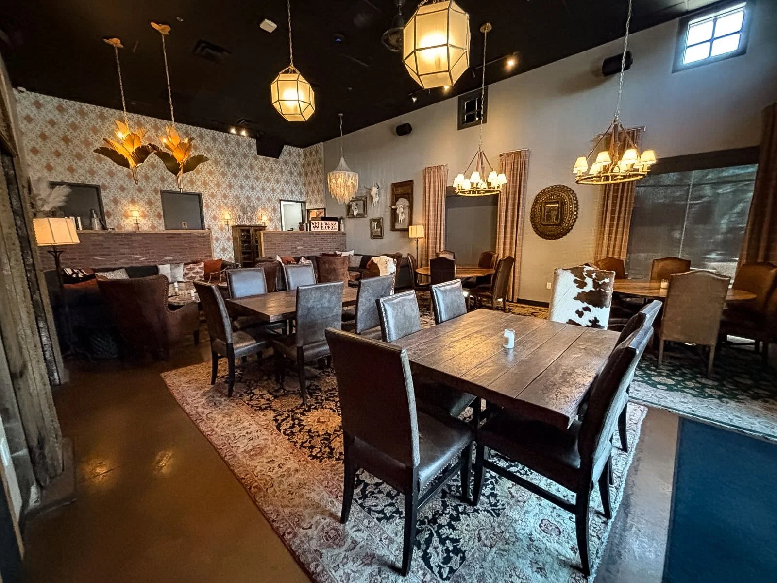 Interior of a cozy restaurant with diverse seating including a large central wooden table with black leather chairs, and plush armchairs along the wall. The space features soft lighting from chandeliers and wall lamps, patterned wallpaper, and area rugs, creating an inviting atmosphere.