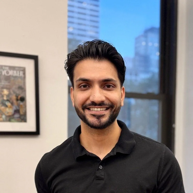 A smiling man with dark hair and a beard in a black polo shirt, indoors, with large windows showing city buildings in the background.