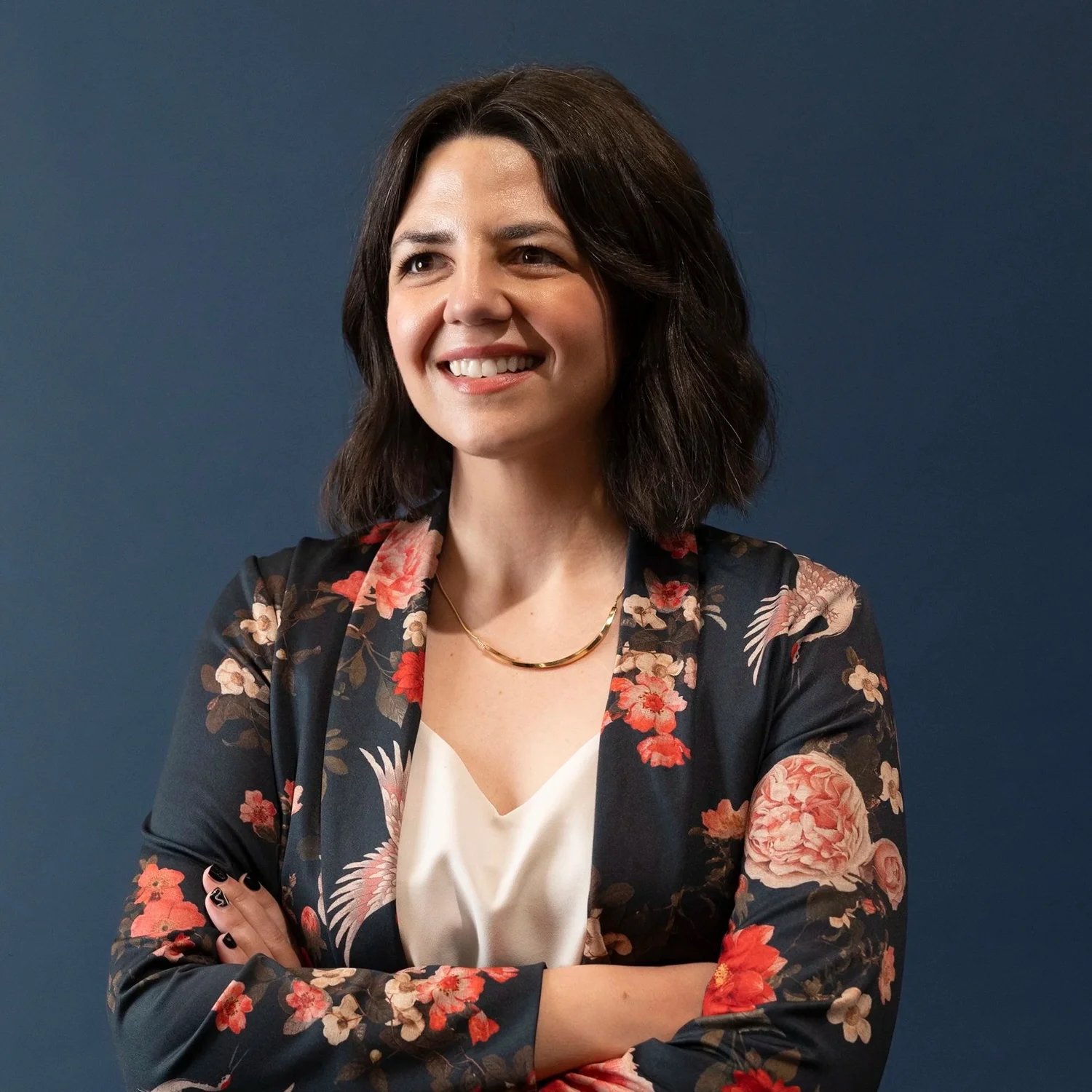 A woman with shoulder-length dark hair, smiling, wearing a floral blazer over a white top, with a gold necklace, in front of a dark blue background.