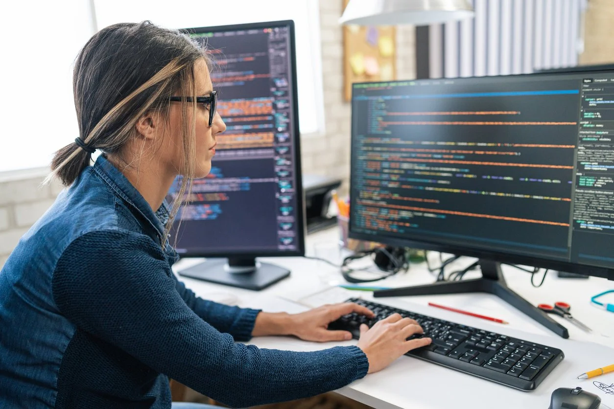 Woman working on coding or programming at a desk with dual monitors displaying code and data.