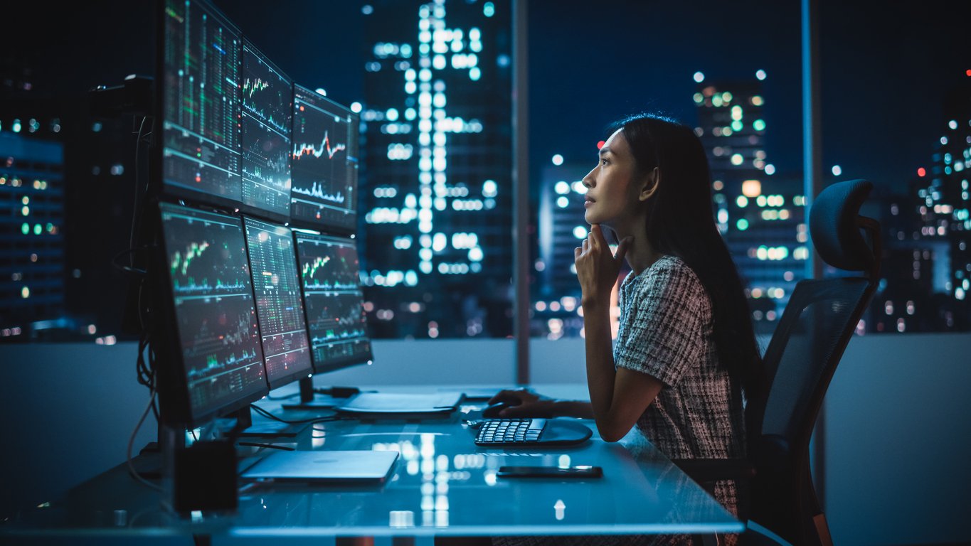 A woman working at a desk with four computer monitors displaying stock market graphs in a high-rise office at night, overlooking a cityscape.