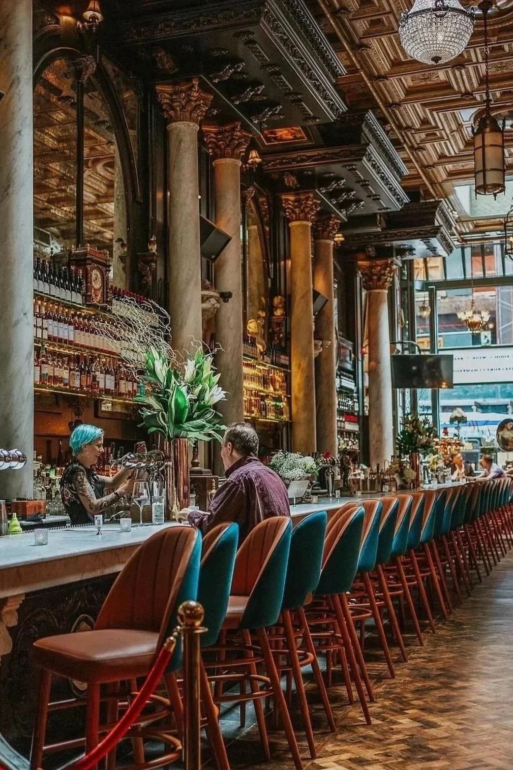 Elegant bar interior with ornate woodwork and tall columns, a long marble counter with colorful chairs, a bartender serving drinks, and large floral arrangements.