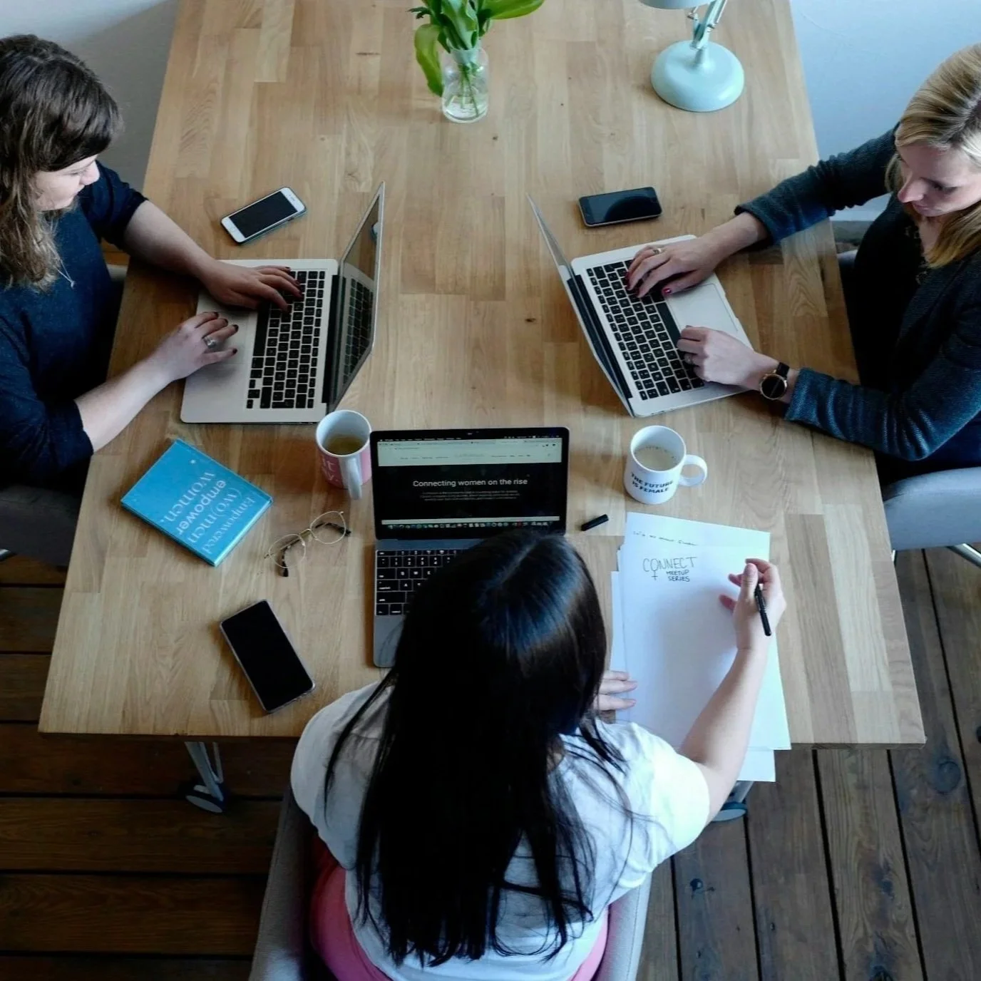 Top-down view of four women working together at a wooden table with laptops, notebooks, coffee mugs, and smartphones.
