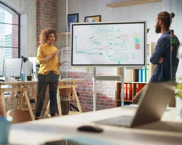 Two people in a modern office, one woman presenting a chart on a whiteboard while the man listens, with laptops and office supplies around.