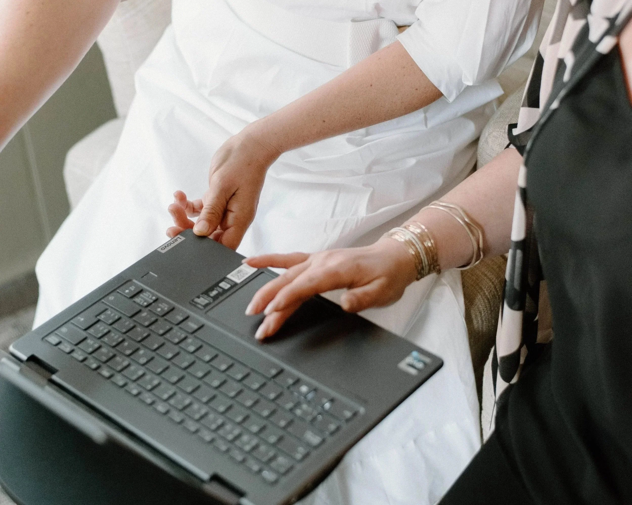 Person in hospital gown sitting on bed working on a laptop with a healthcare worker nearby.