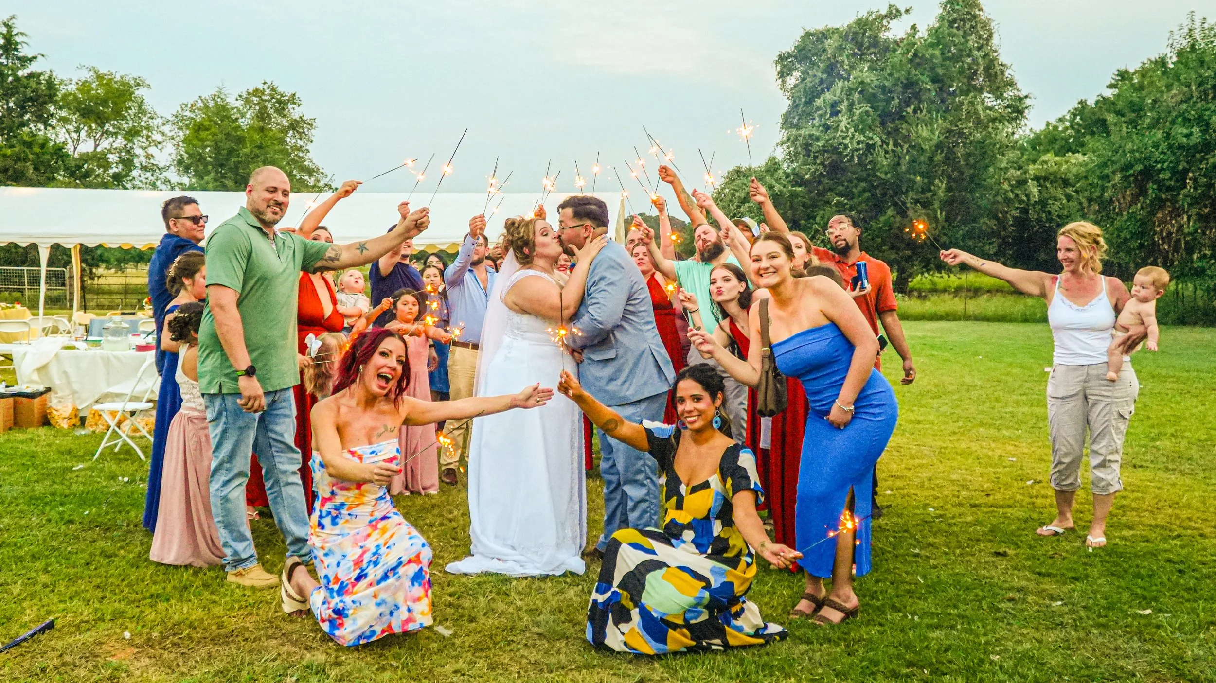Group of people celebrating a wedding outdoors, including the bride and groom kissing in the center while holding sparklers, surrounded by friends and family holding sparklers and smiling.