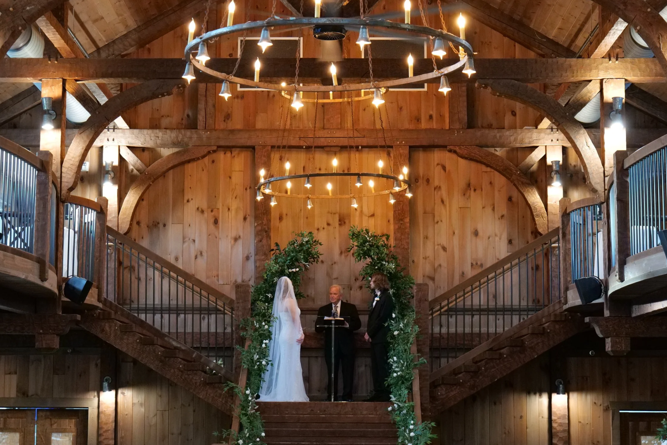A wedding ceremony taking place inside a rustic wooden venue. A bride and groom stand face to face before an officiant, under a circular chandelier hanging from a wood-beamed ceiling, with stairs and greenery in the background.