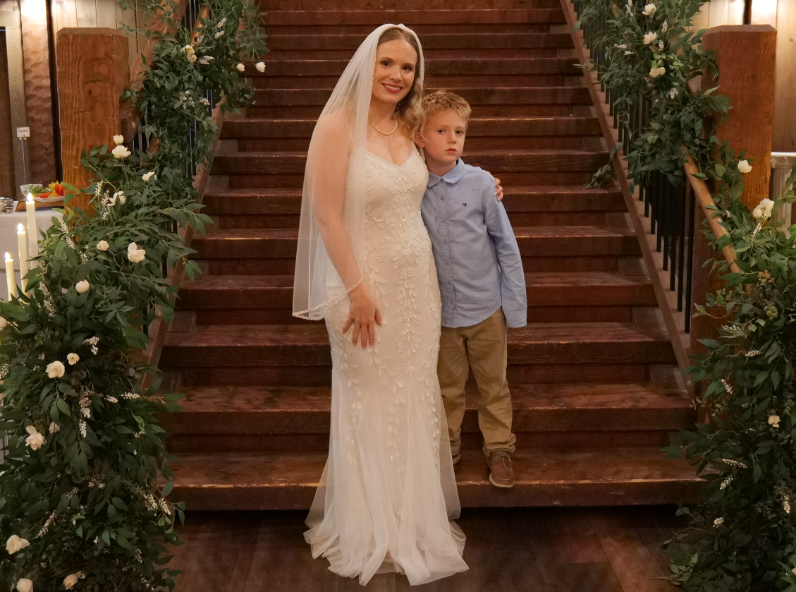 A woman in a wedding dress and veil standing on a wooden staircase next to a young boy in a light blue shirt and khaki pants, with floral decorations on the staircase railing.