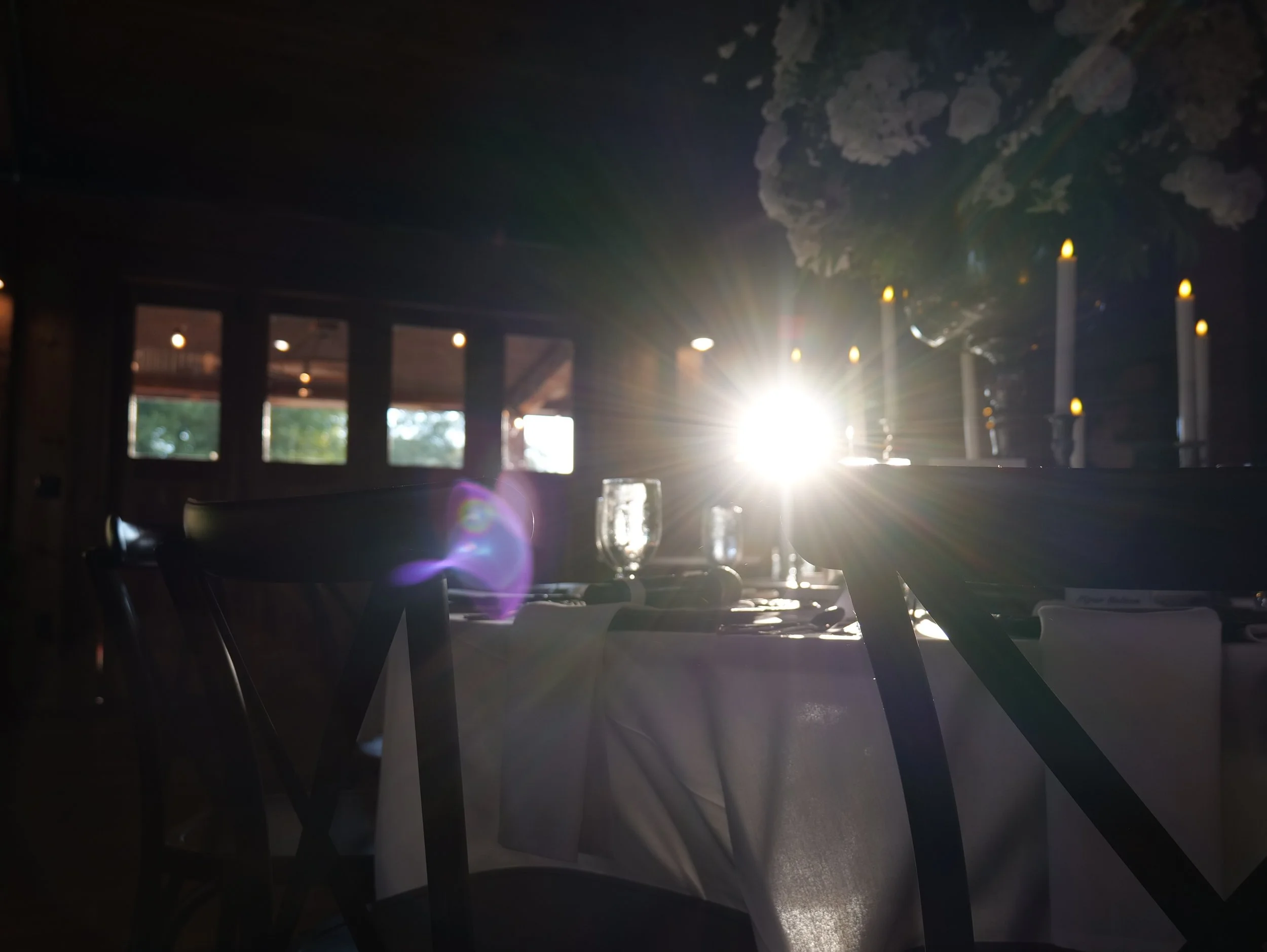 A table set with glassware and candles, illuminated by sunlight streaming through windows, with floral arrangements and a rustic interior.