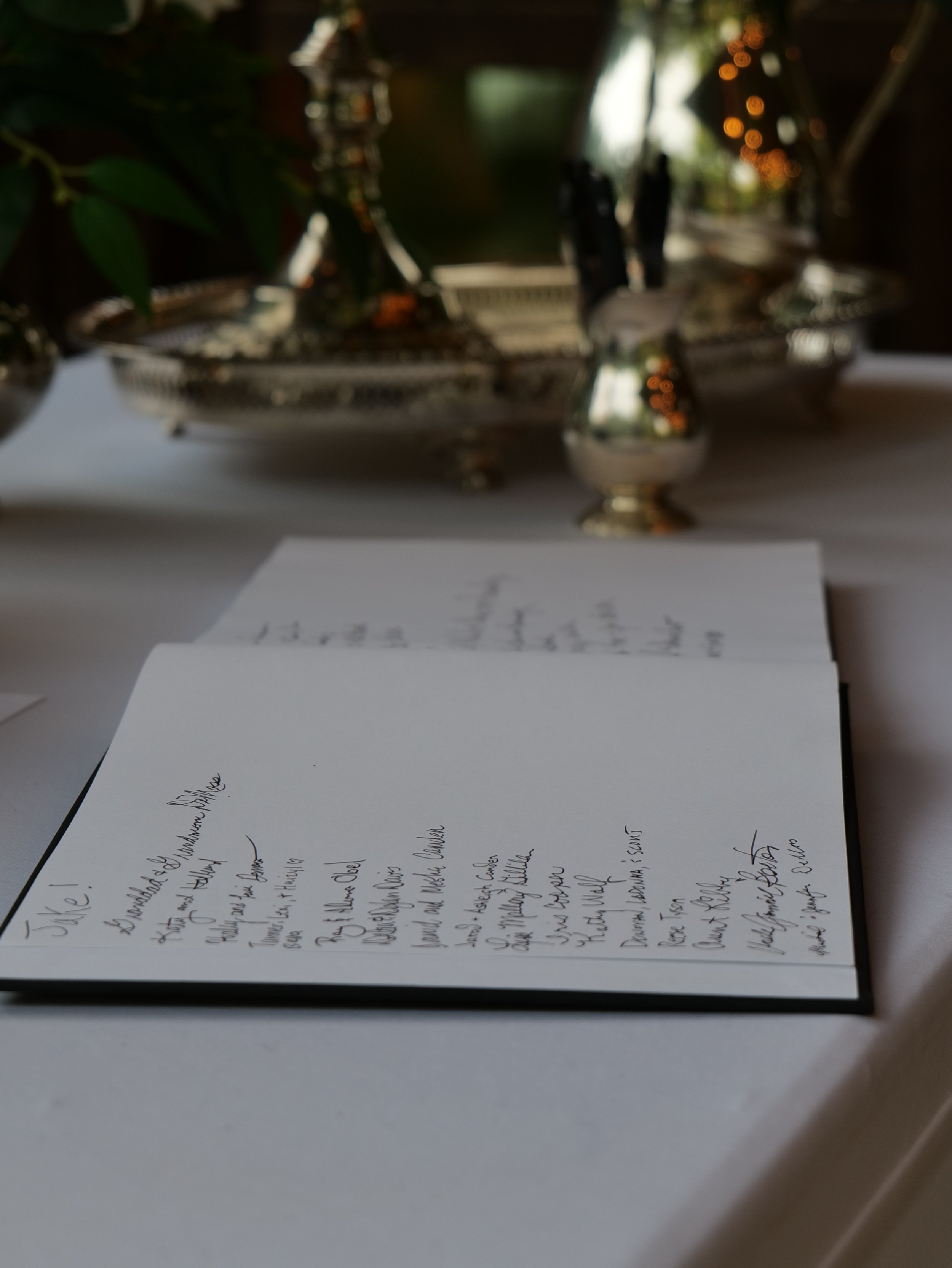 Close-up of a handwritten menu or note on a table, with a silver tray or dish in the background containing plants and decorative objects, in a setting with warm lighting.