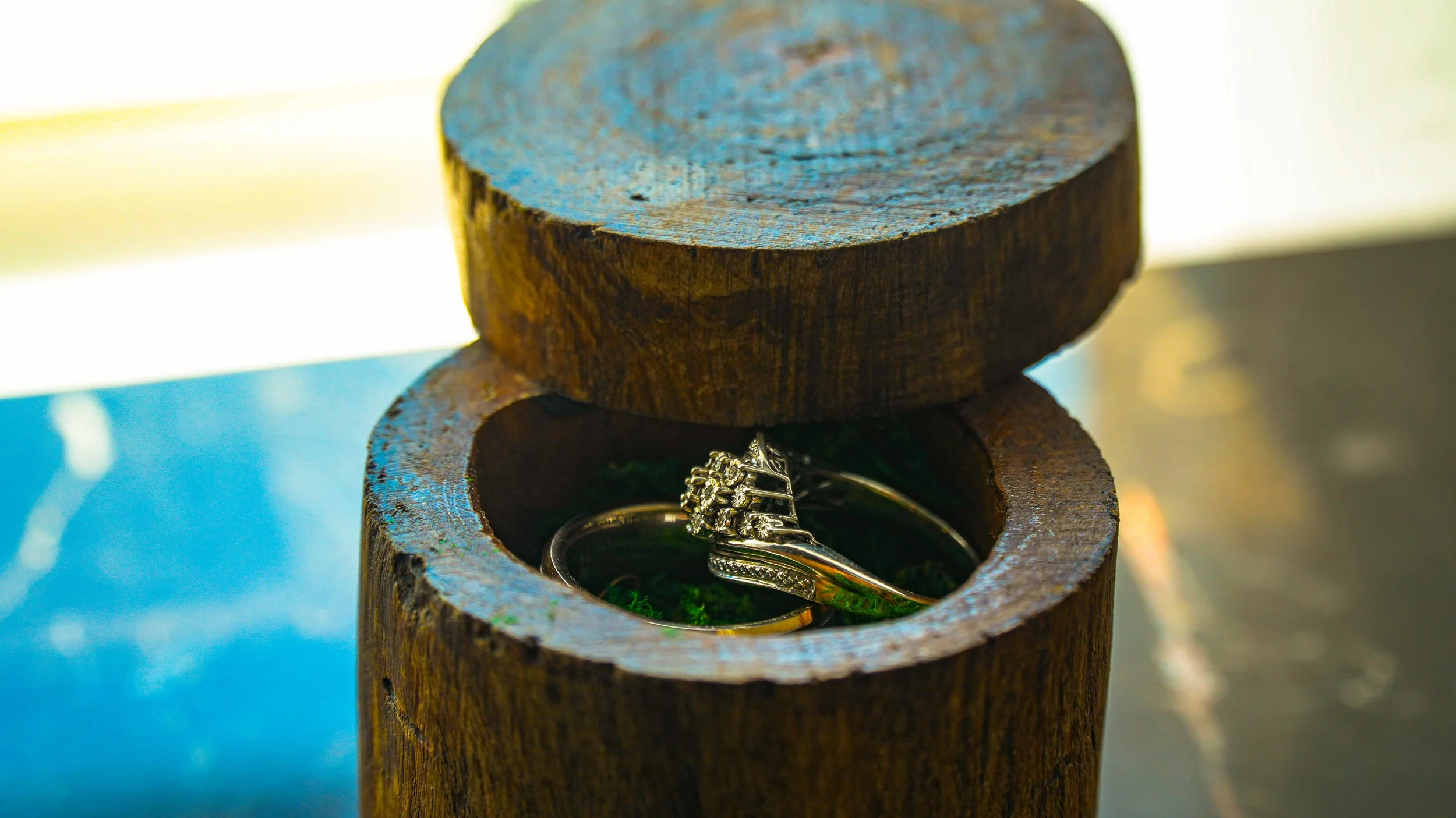 A wooden ring box with a silver ring inside, resting on a wooden surface with sunlight in the background.