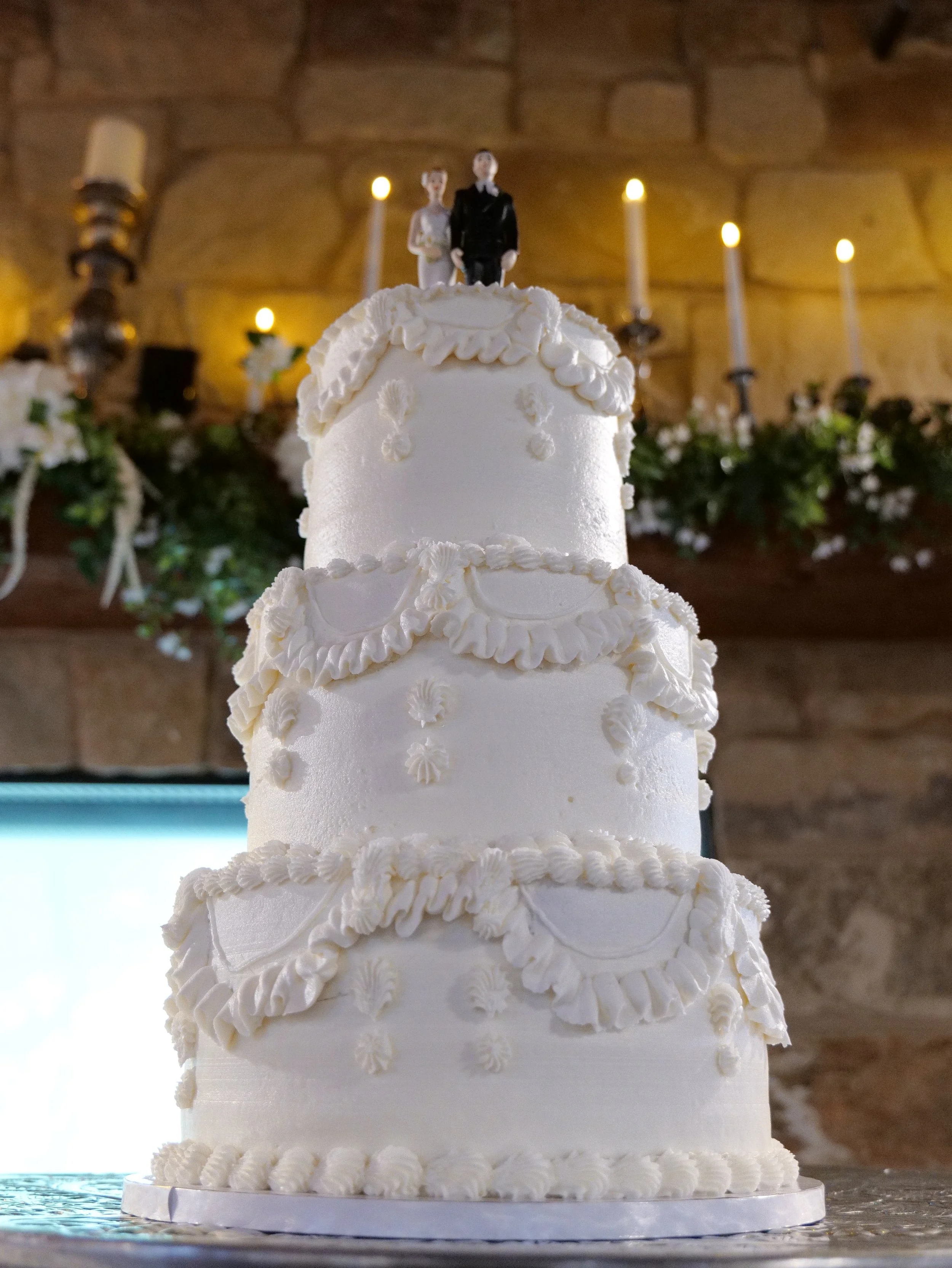 A three-tier white wedding cake with elaborate piped icing decorations, topped with a bride and groom figurine, in a warmly lit room with candles and floral decor in the background.