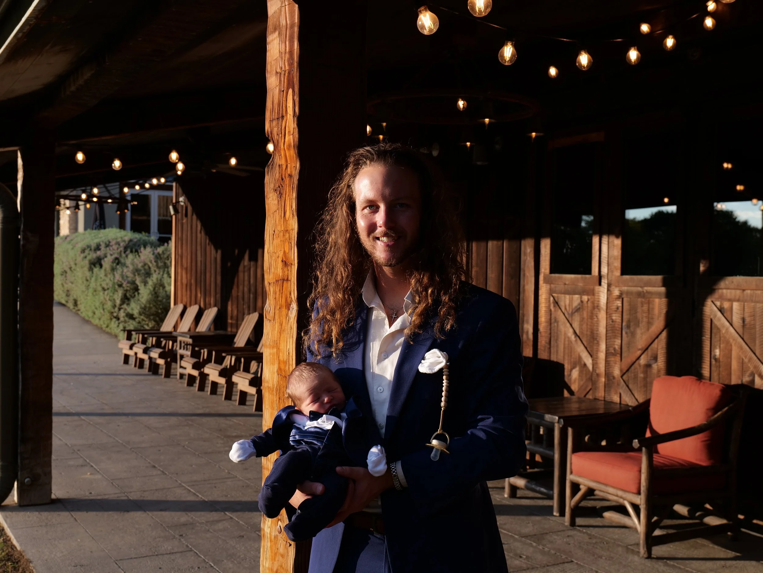 A man with long curly hair and a beard holding a newborn baby outside under string lights, in front of a wooden building with patio furniture.