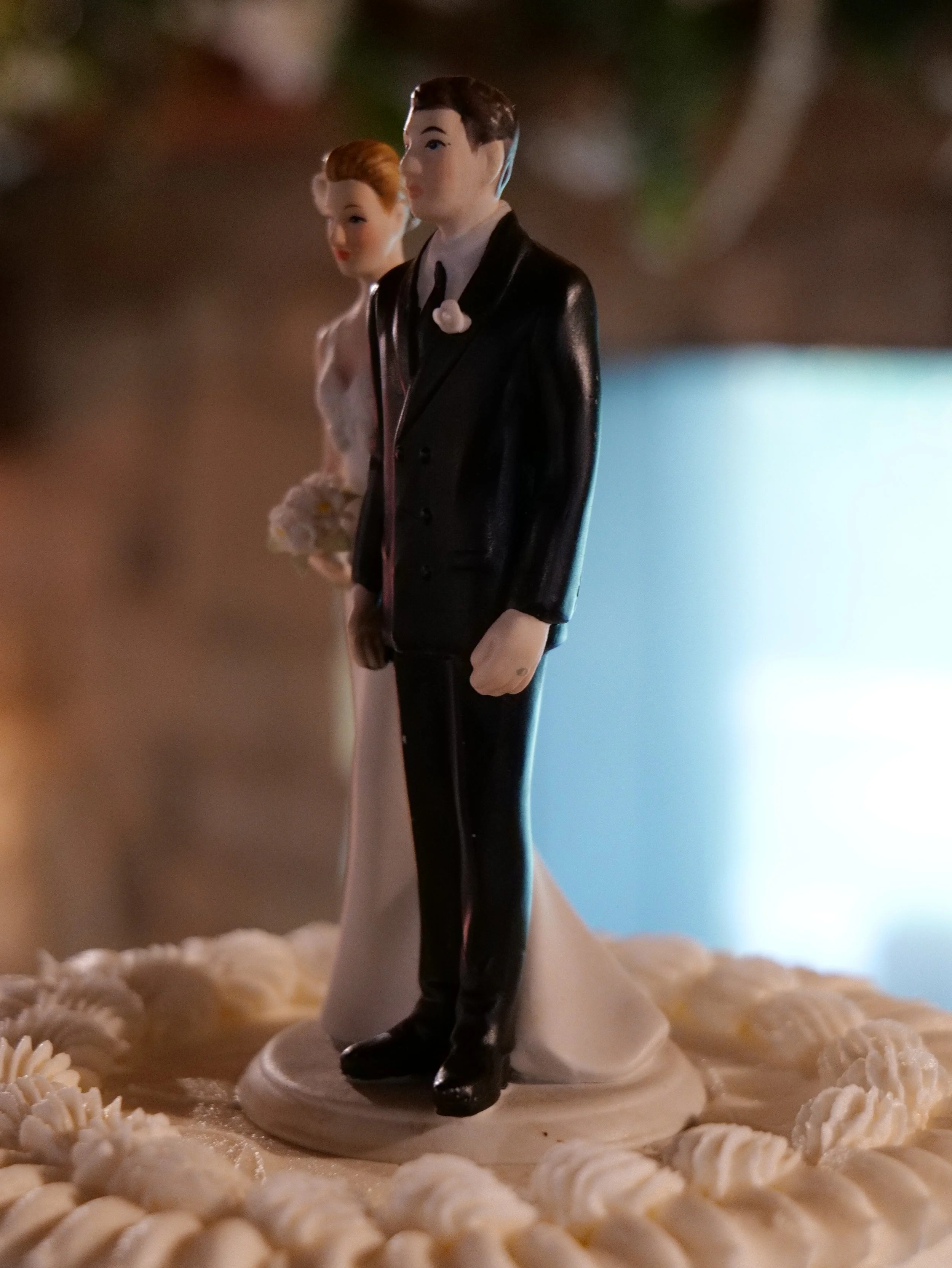 A figurine of a bride and groom on a wedding cake, with the groom dressed in a black tuxedo and the bride in a white wedding gown, holding a bouquet.