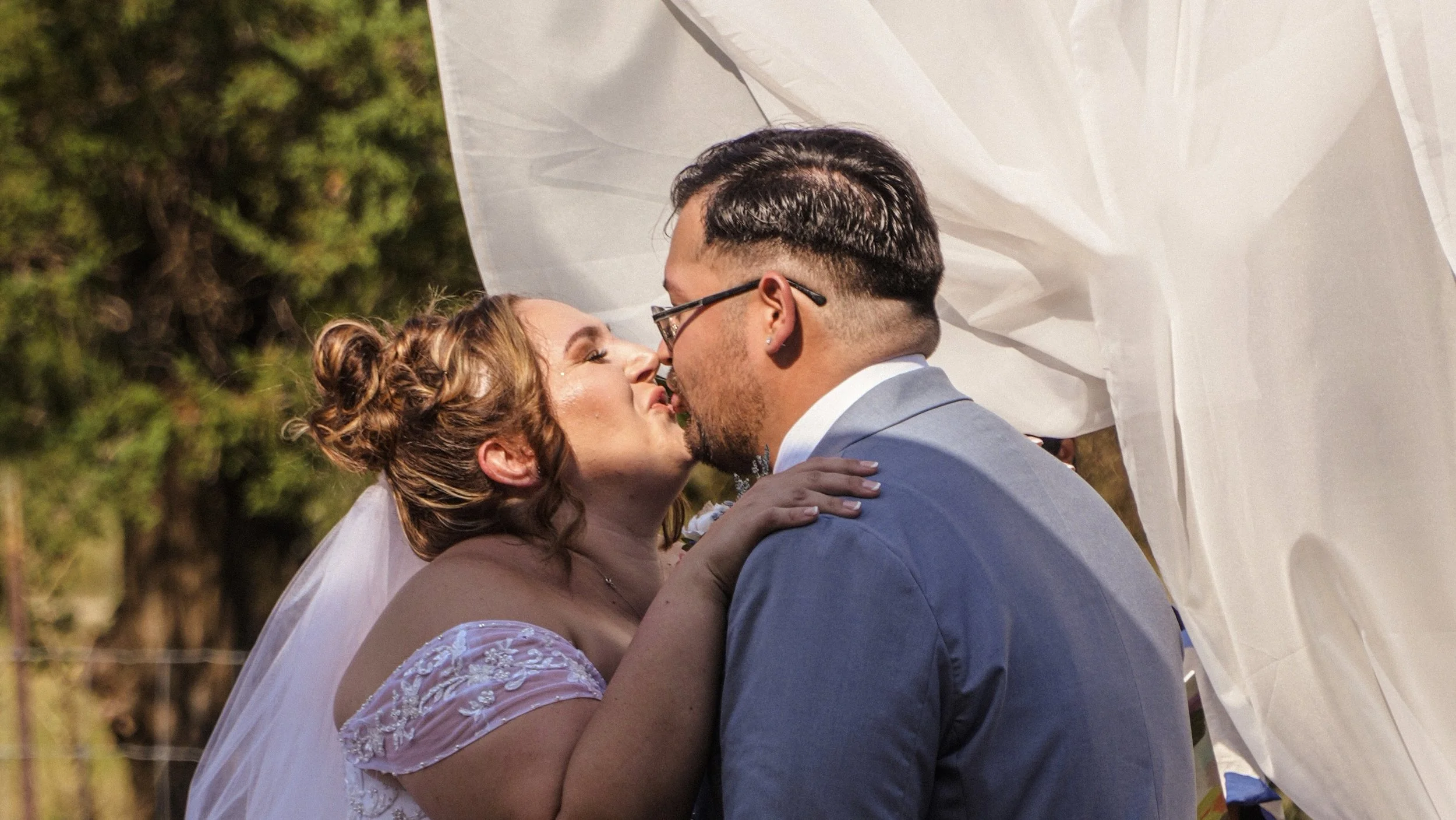A bride and groom kissing outdoors during their wedding, with trees in the background and white fabric above them.