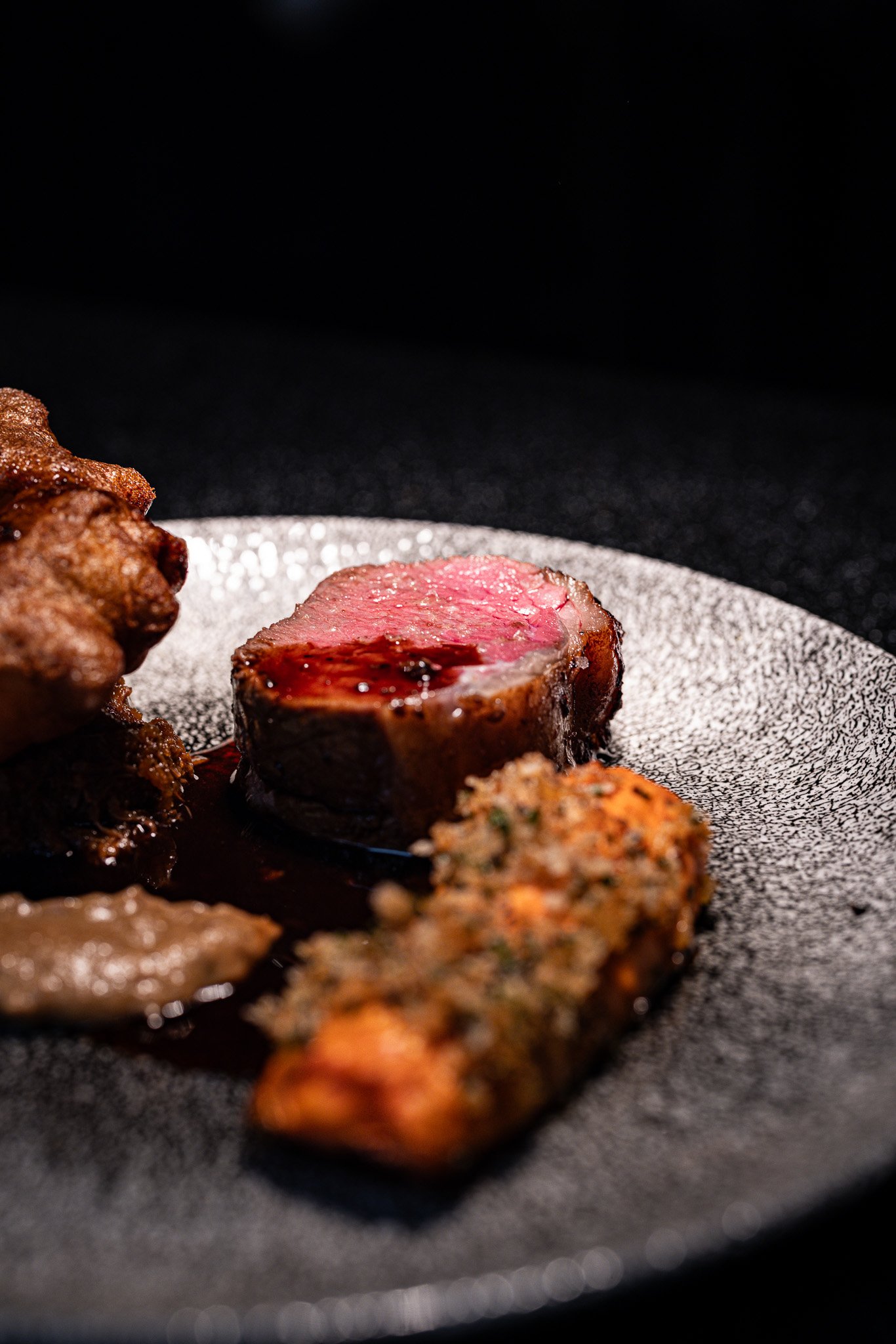 Close-up of a piece of cooked steak on a black textured plate, with other pieces of meat around it, against a dark background.
