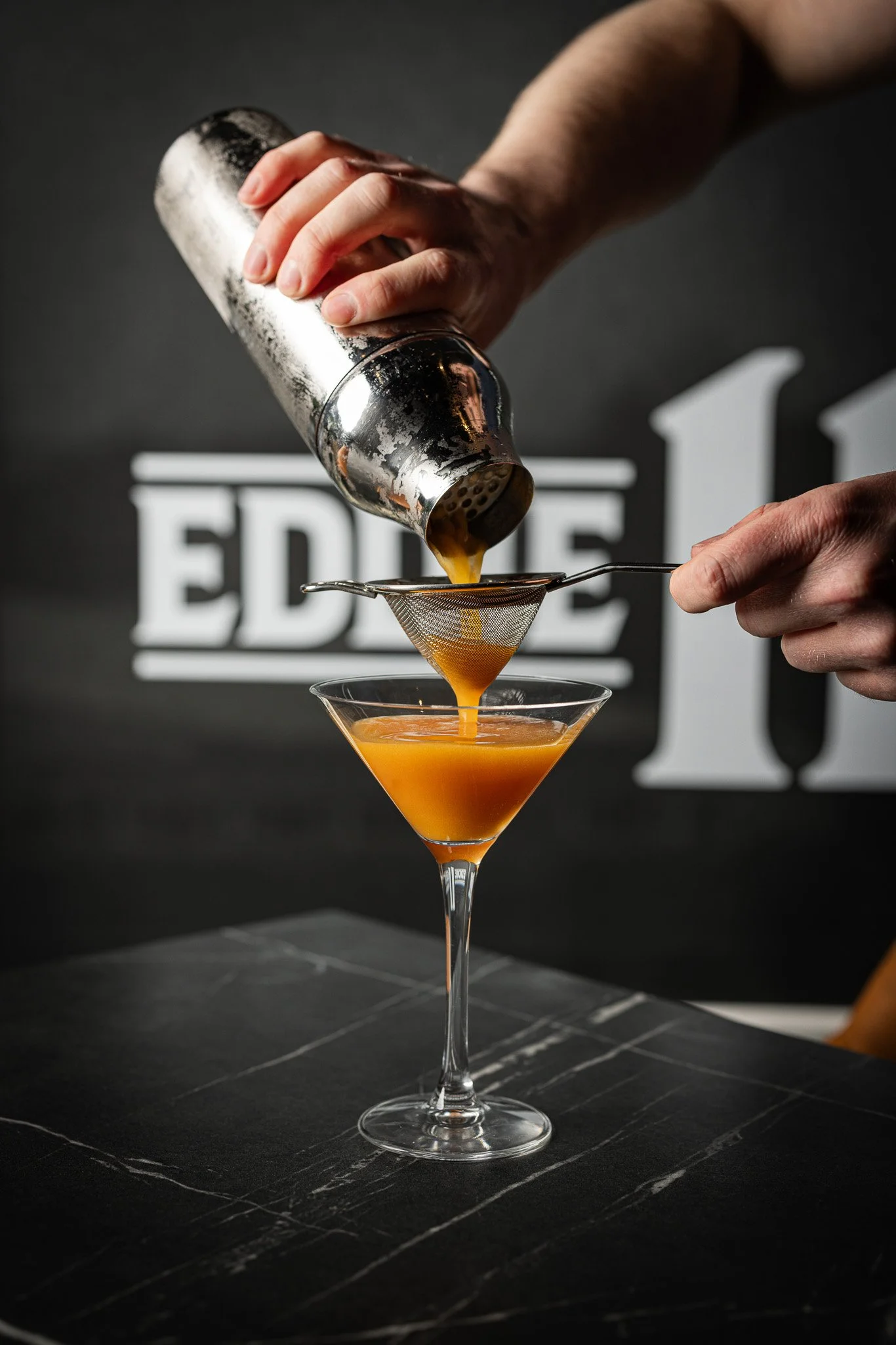 A bartender pouring orange liquid through a cocktail strainer into a martini glass, with a black background and the word 'EDO' partially visible.