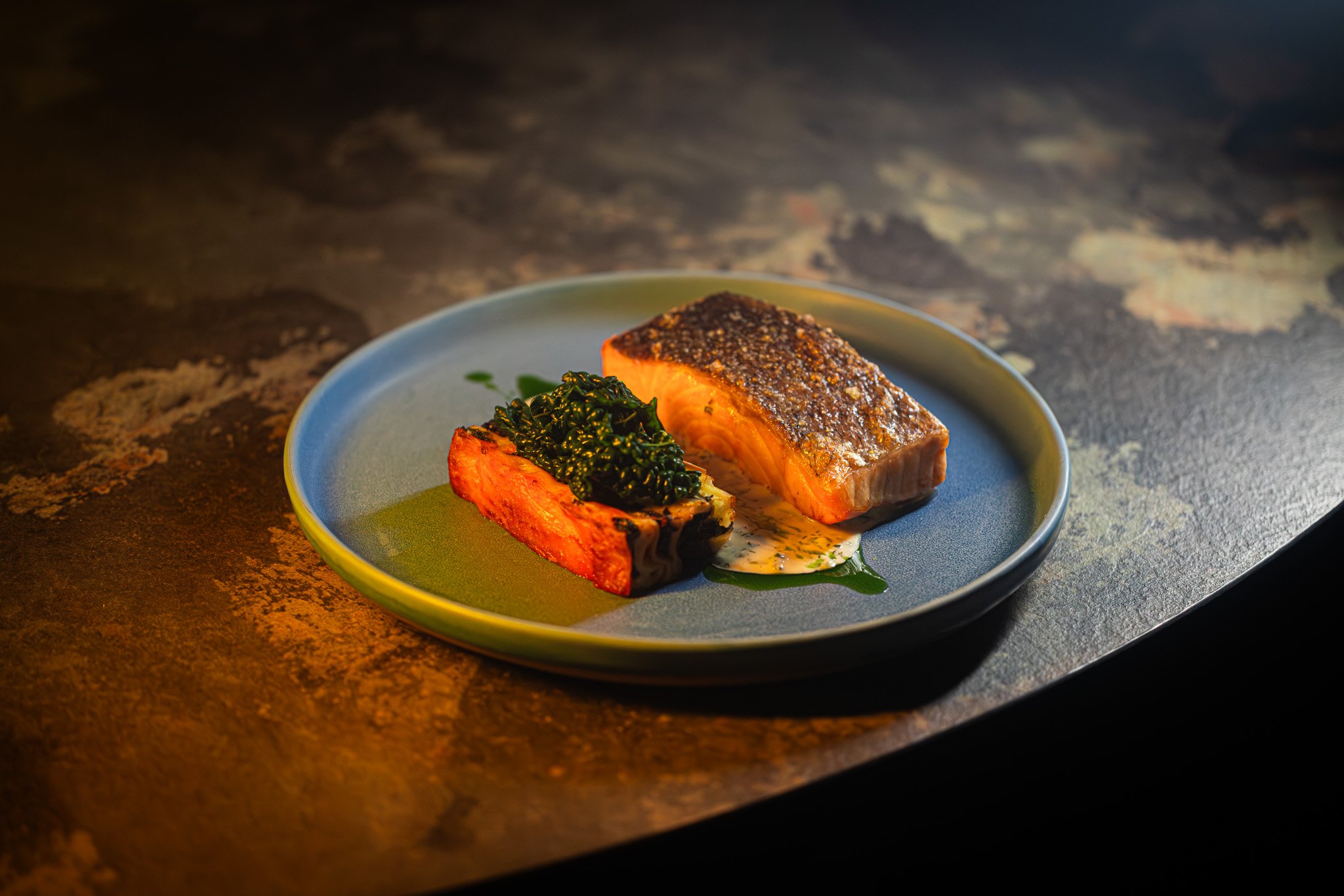 A plate with a cooked fish fillet, a roasted sweet potato topped with greens, and a green sauce on the side, on a textured dark surface.