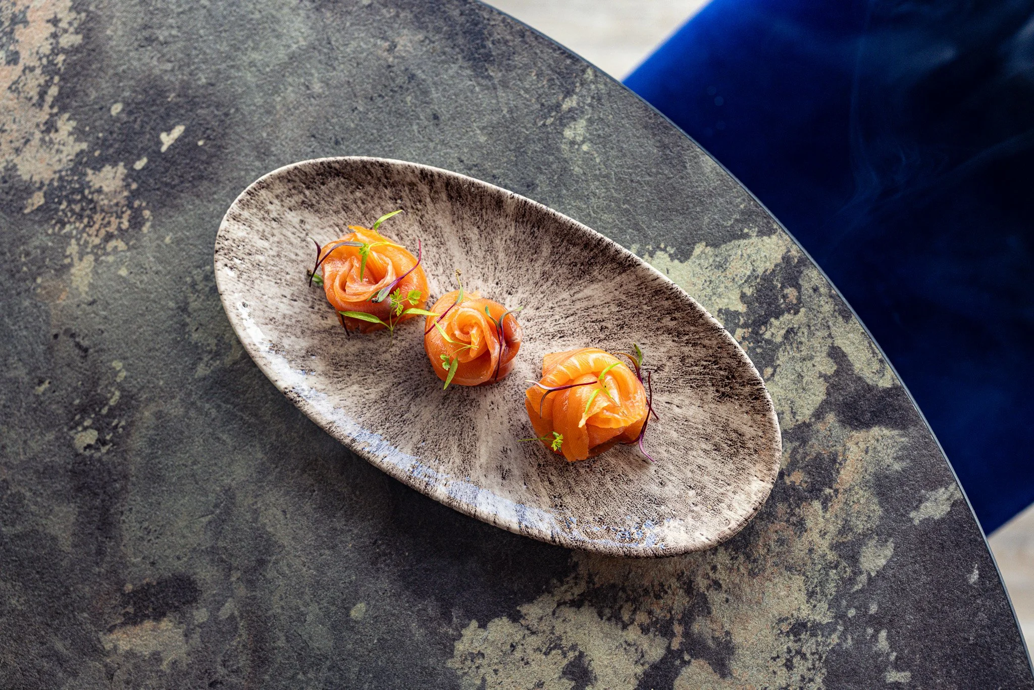 Three salmon proscuitto rose appetizers garnished with microgreens on an oval ceramic plate, placed on a dark textured table with a blue chair partially visible.