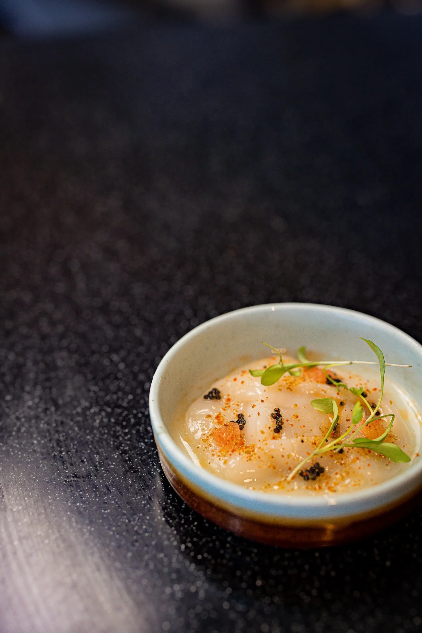 A small dish with a gourmet appetizer, garnished with microgreens and specks of black caviar on a dark surface.