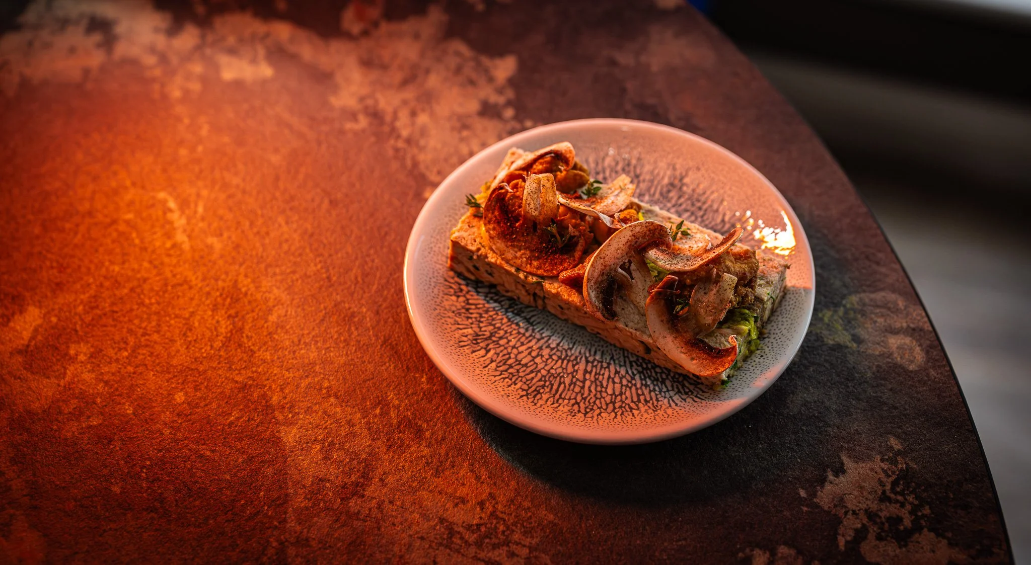A plate of toast topped with sautéed mushrooms, herbs, and seasoning on a textured ceramic plate, on a rustic, dark, reddish-brown table.