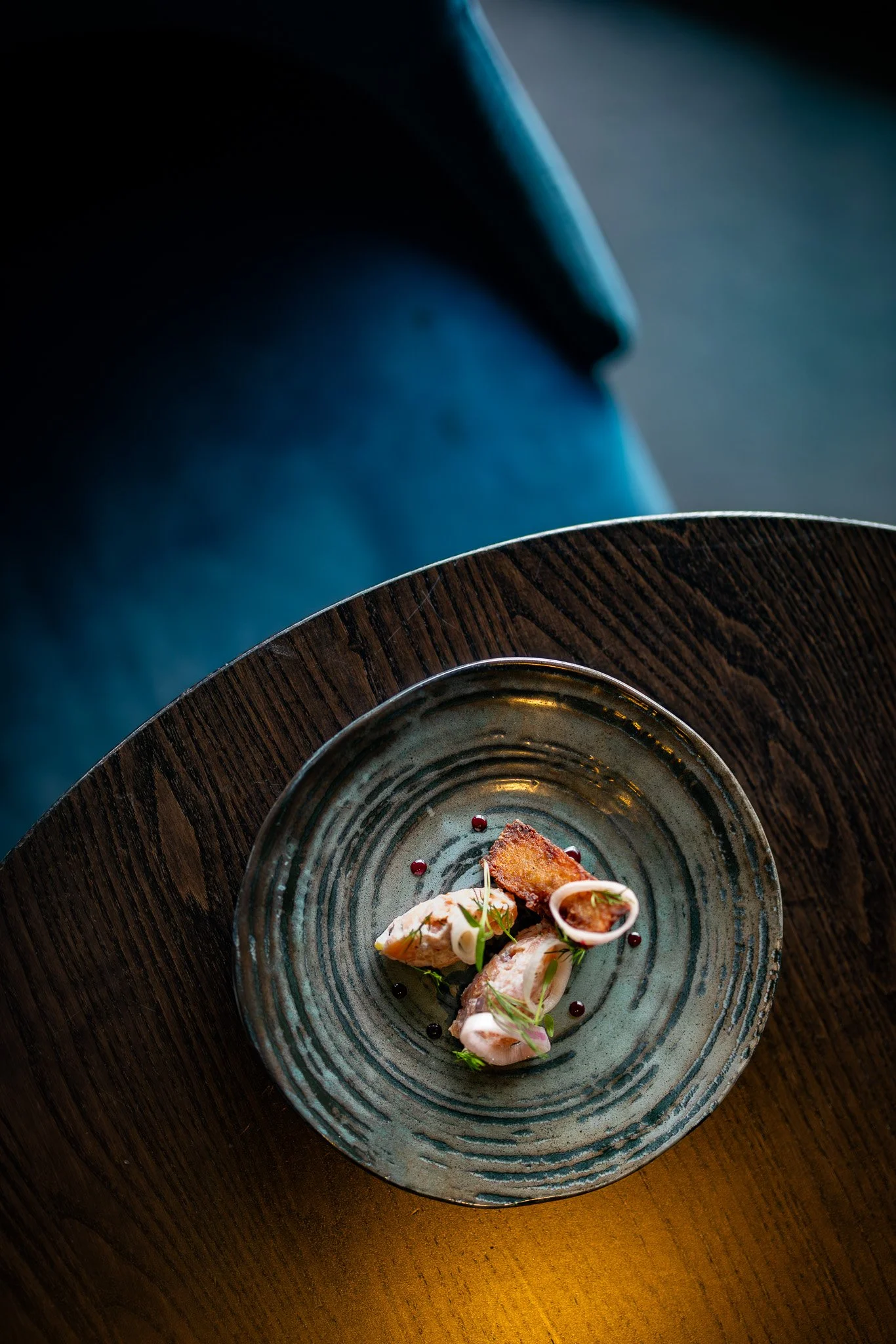 A small plate of gourmet food, including small pieces of fish or meat, garnished with herbs, served on a rustic ceramic plate with dark accents, placed on a wooden table.