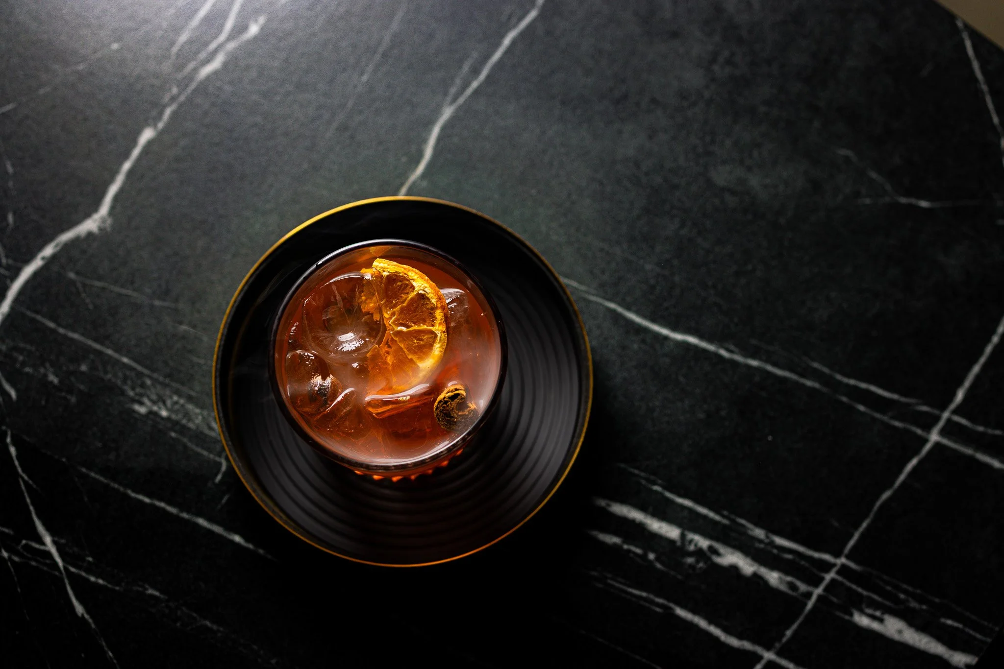 A glass of iced tea with a lemon slice and ice cubes, placed on a black plate with a gold rim on a black marble surface.