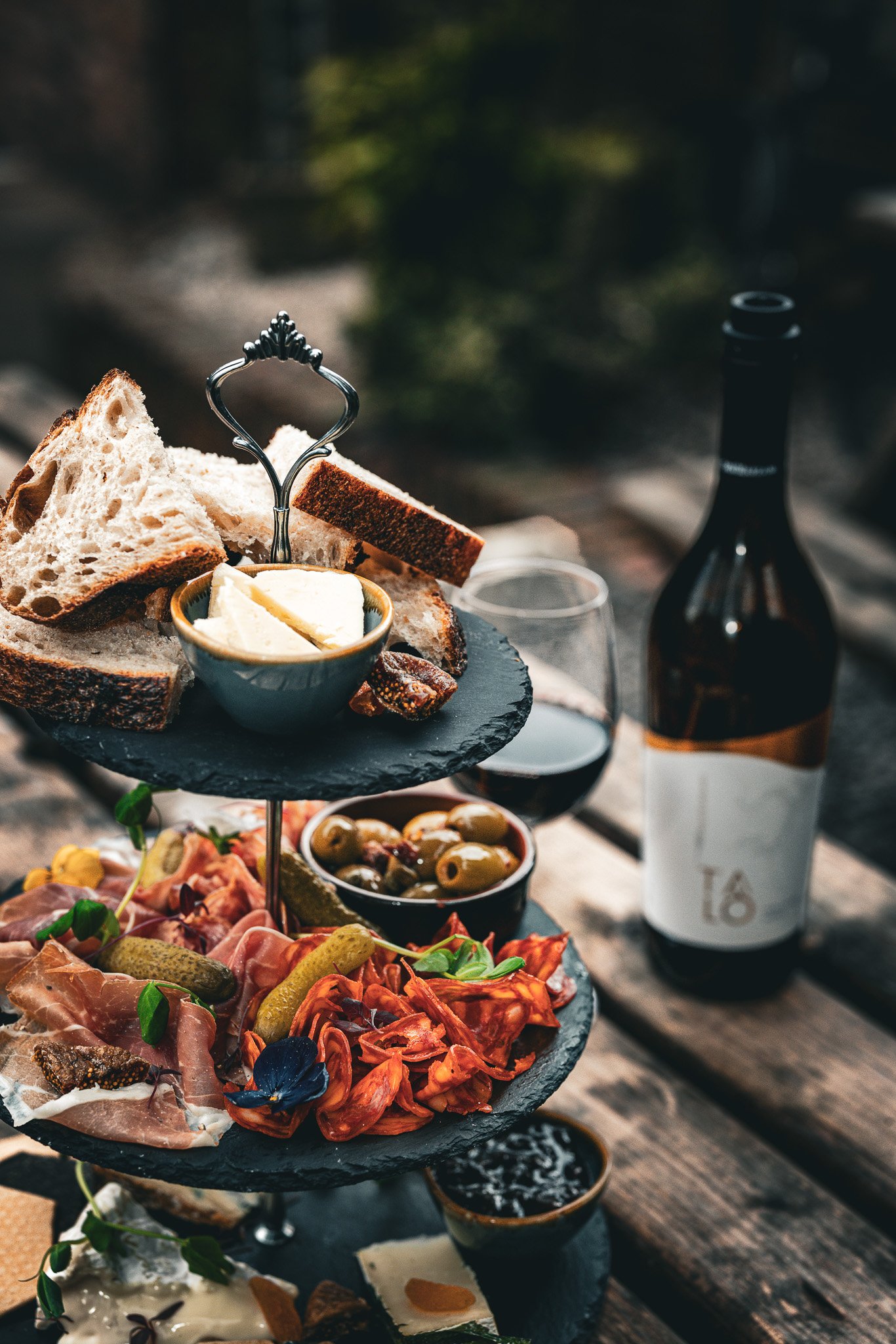 A three-tiered black slate serving platter with bread, cheese, olives, cured meats, pickles, and a blue edible flower, set on a rustic wooden table with a bottle of red wine and a glass in the background.