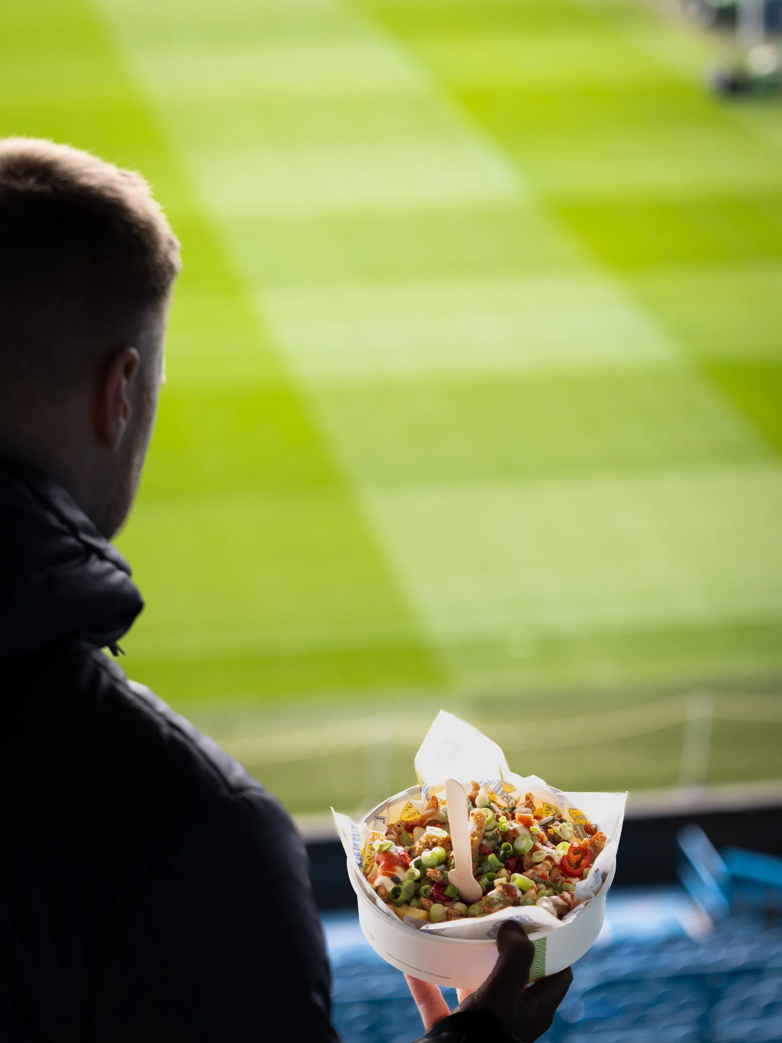 A man holding a bowl of loaded fries with various toppings while looking out onto a green field or stadium.