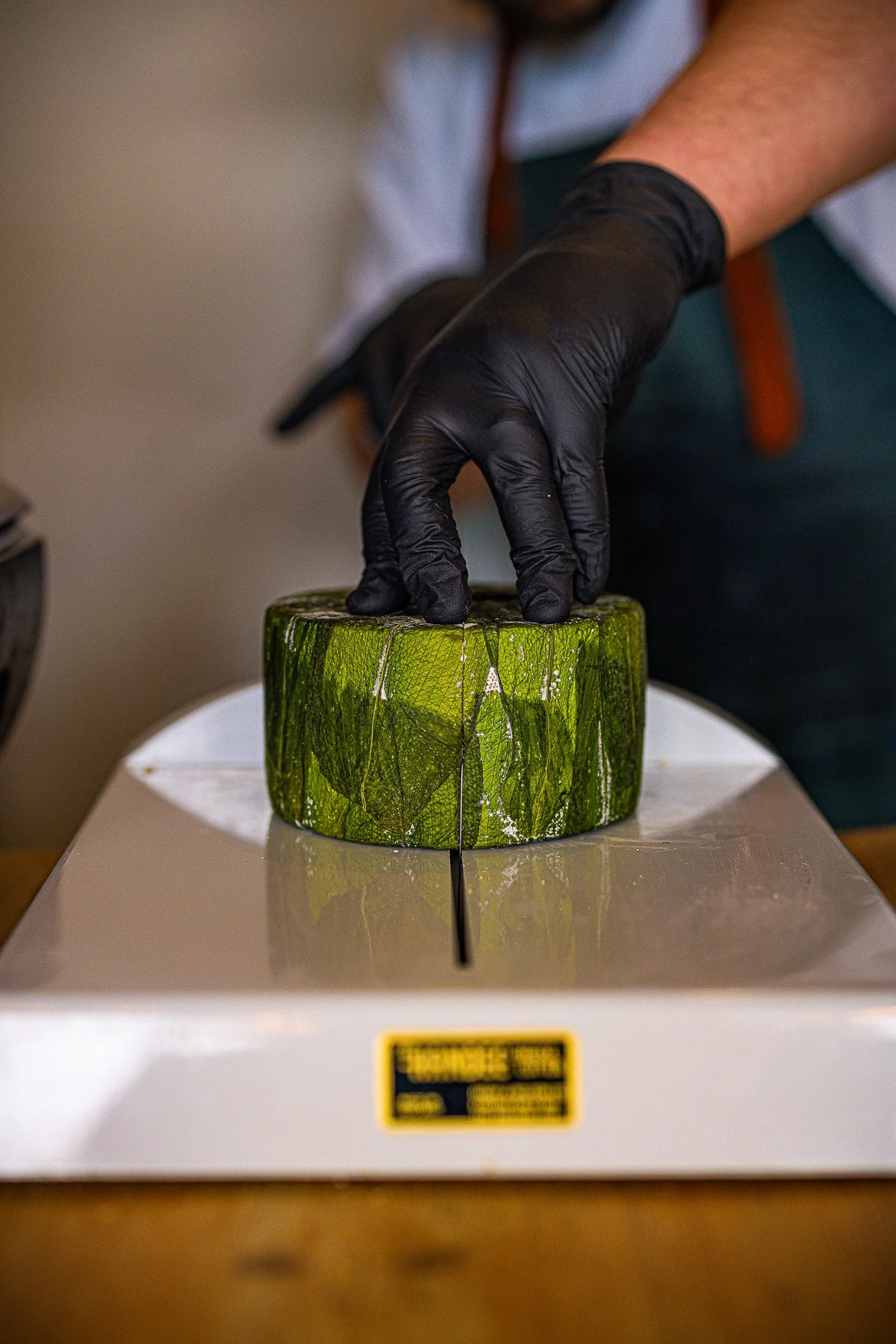A person wearing black gloves is slicing a large green piece of cheese on a white cutting board with a slicer.