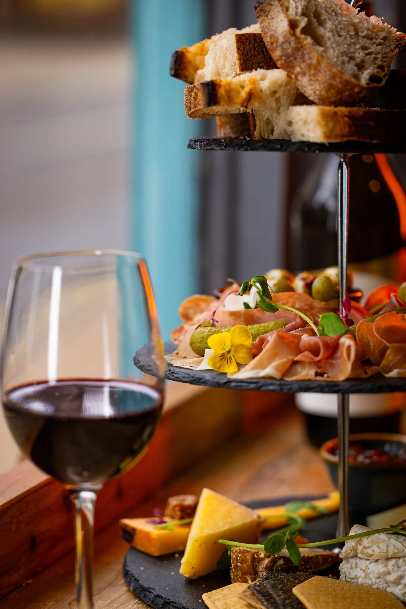 A tiered platter of assorted snacks including sliced bread, cured meats, cheese, and olives, accompanied by a glass of red wine.