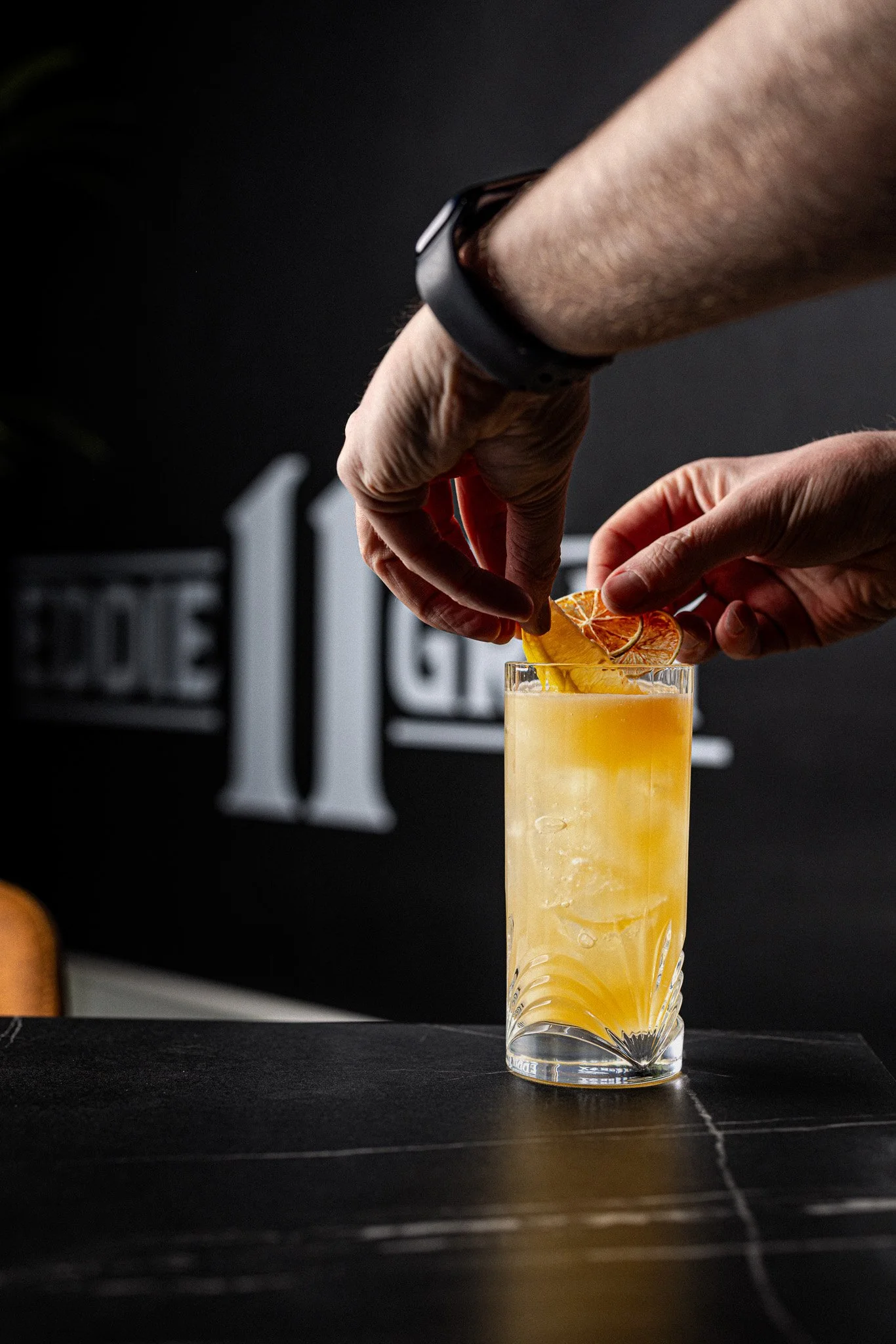 Hands garnishing a yellow cocktail with citrus slices on a black table.