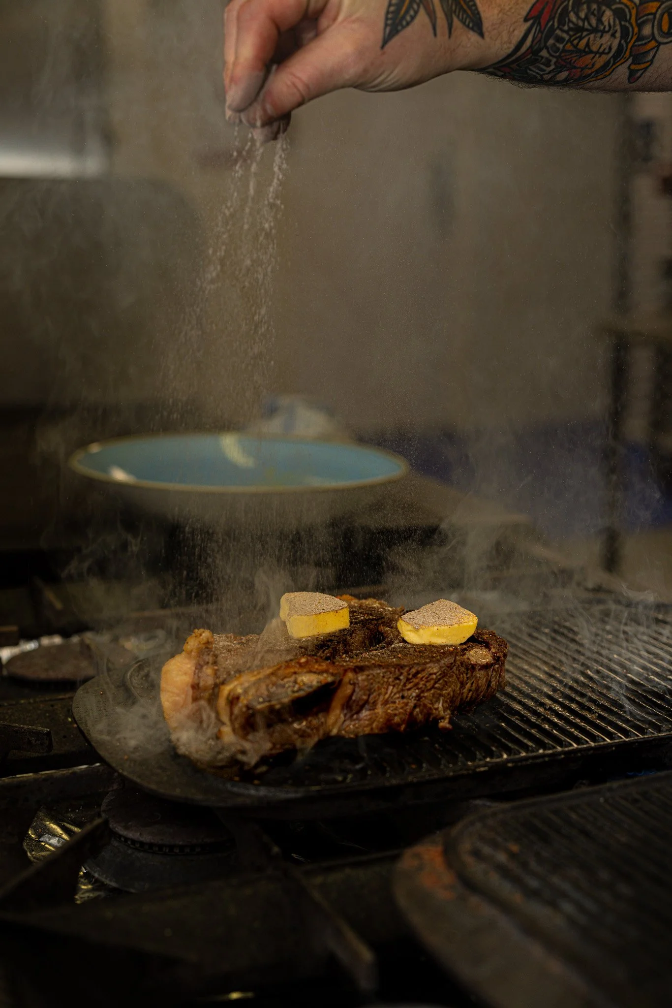 A person seasoning a large steak on a grill with pats of butter melting on top, while black grill marks are visible on the meat.