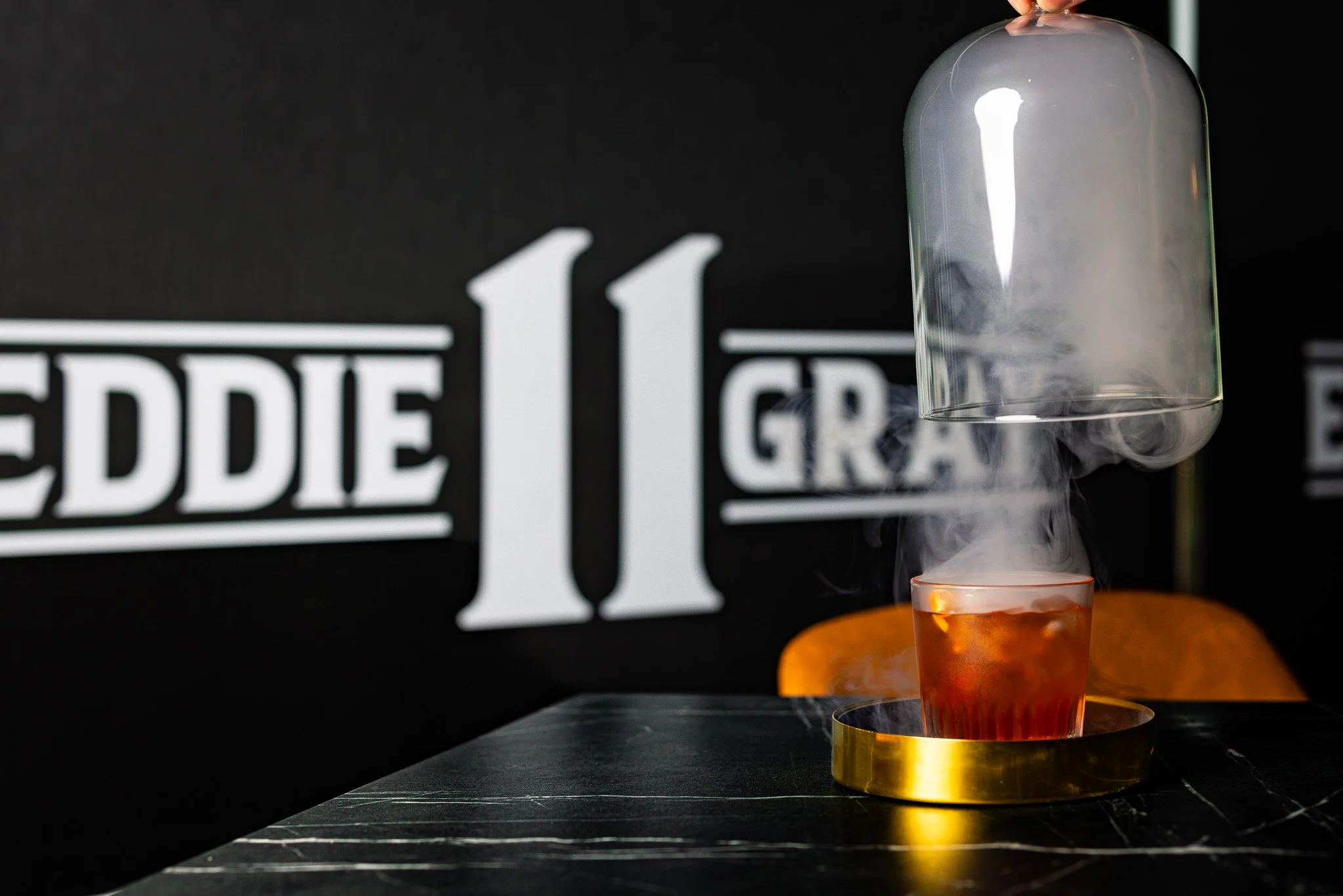 A cocktail glass with a dark red drink has dry ice, creating fog, and is covered by a glass dome, on a black table with a black background and the words 'Eddie 11' in white.