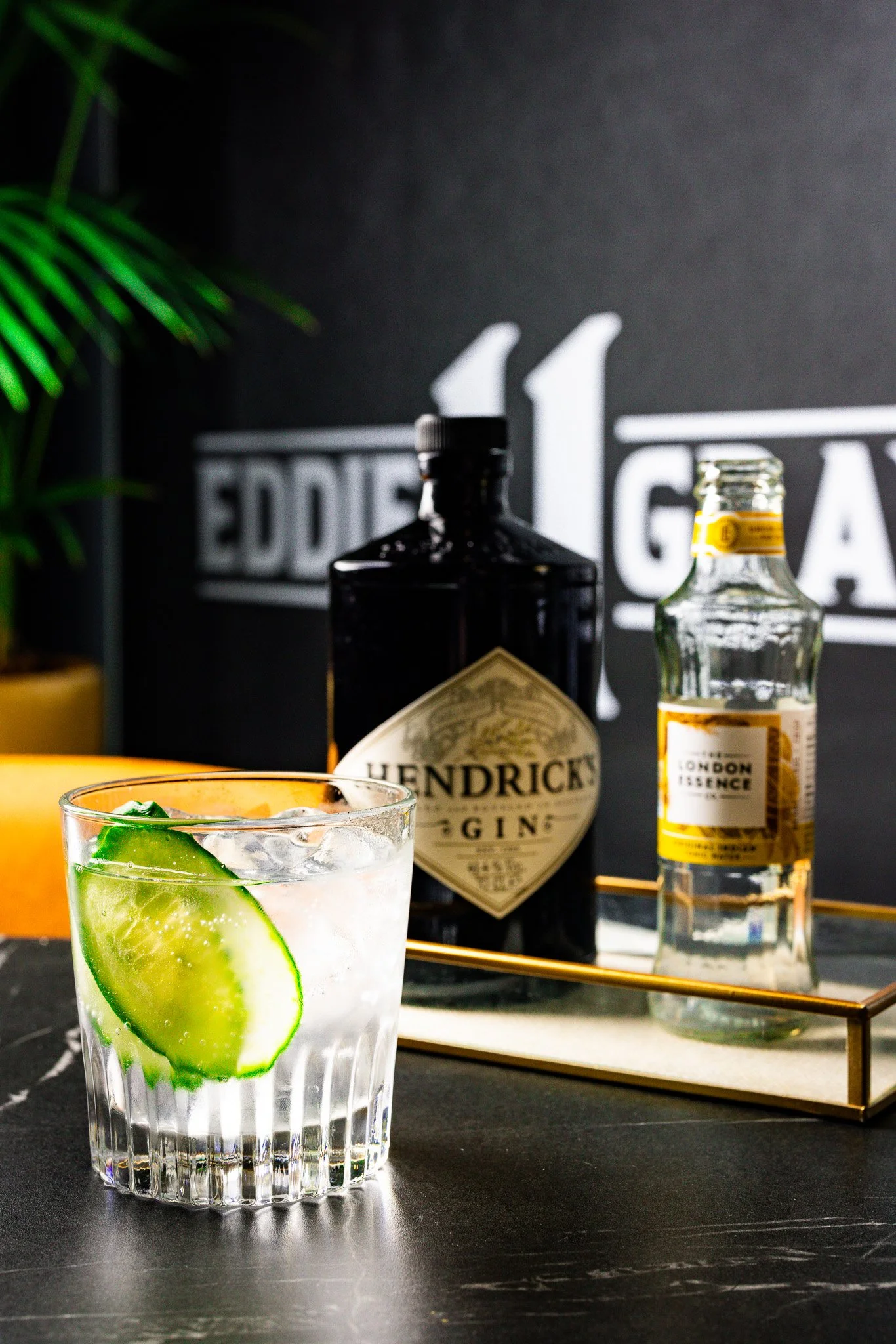 A glass of gin and tonic with cucumber slice on a black surface, with gin and tonic bottles and a blackboard background in a bar setting.