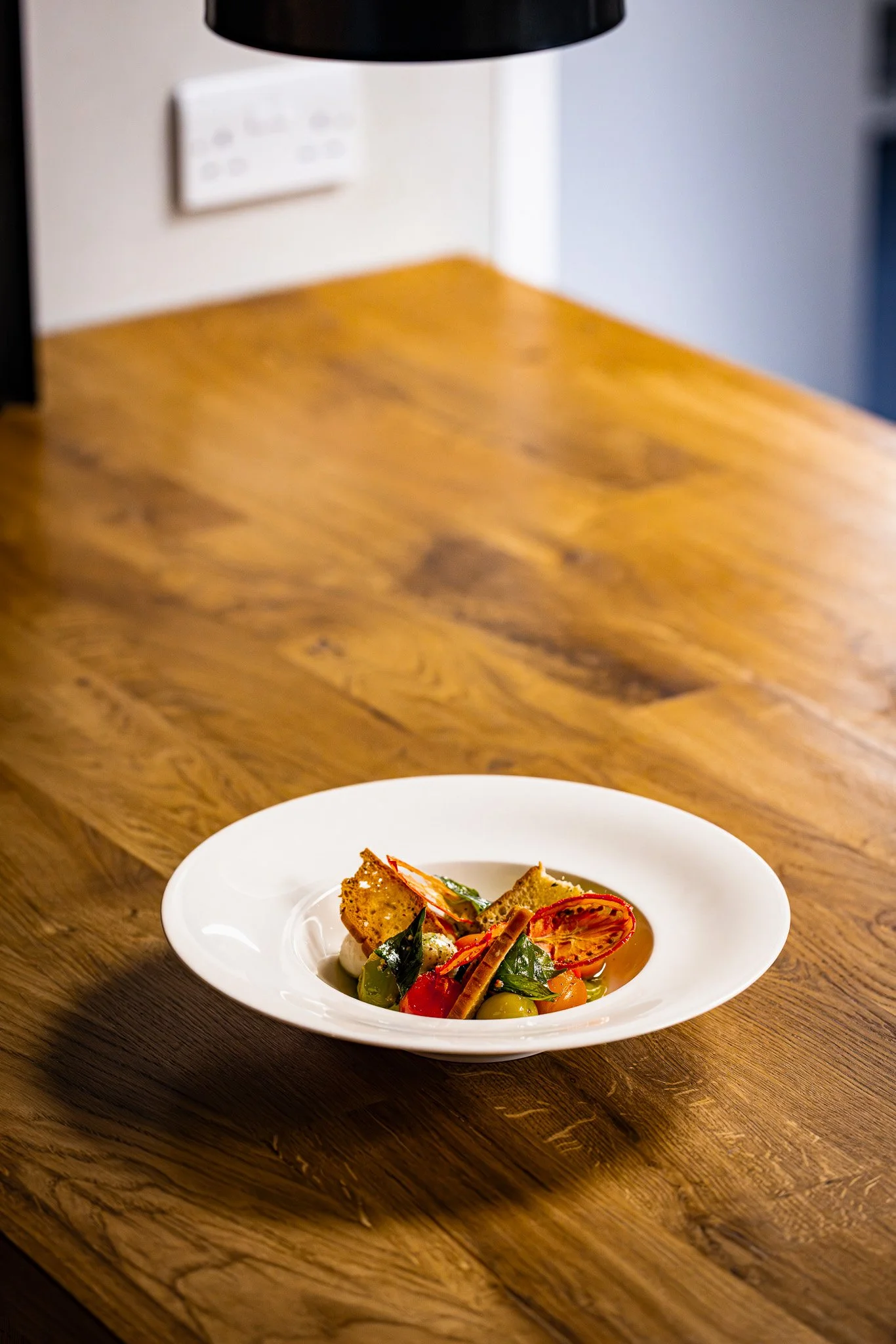 A white plate with a gourmet dish, including vegetables, bread, and garnishes, placed on a wooden table.