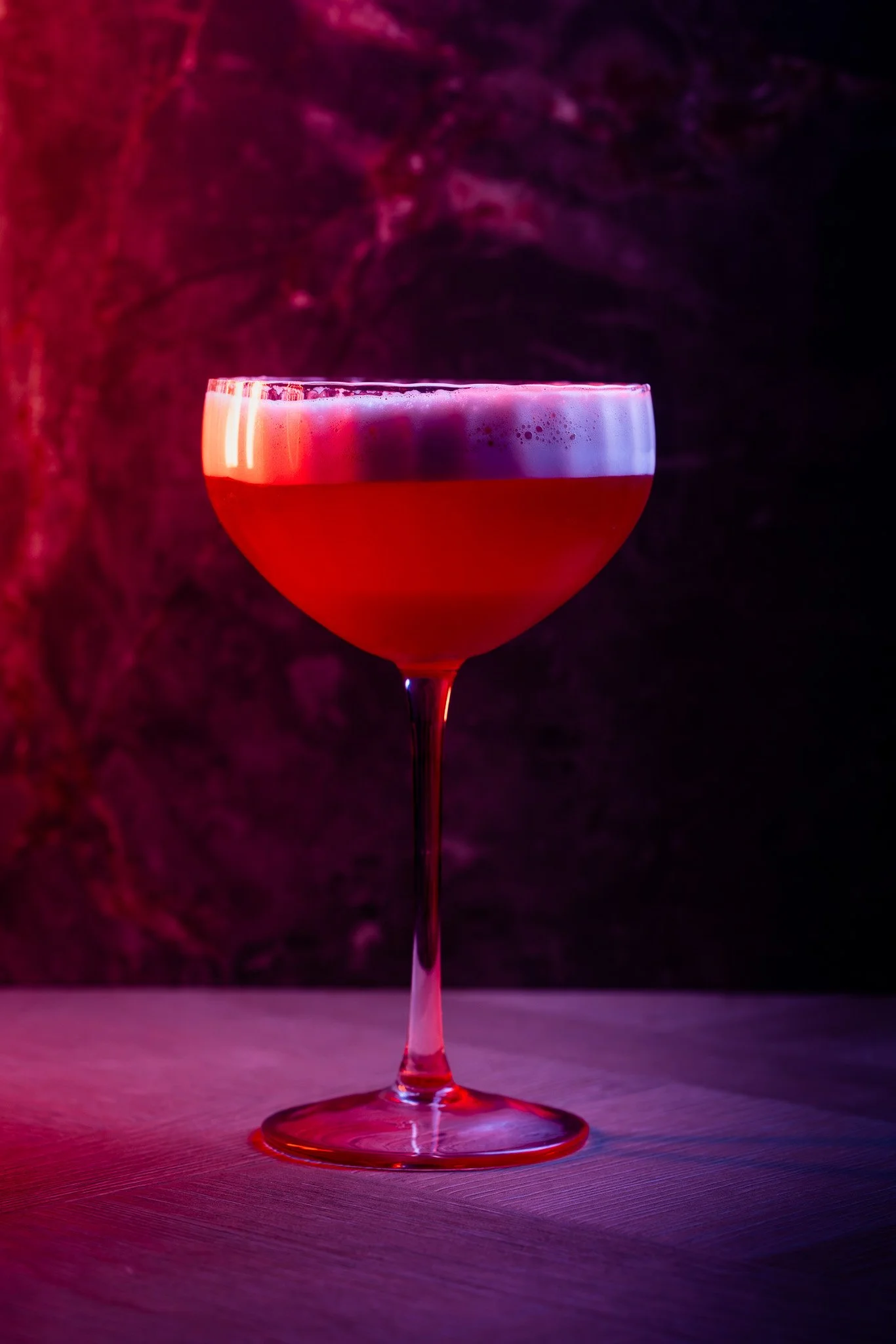 A red cocktail in a coupe glass on a wooden surface with purple and pink lighting in the background.