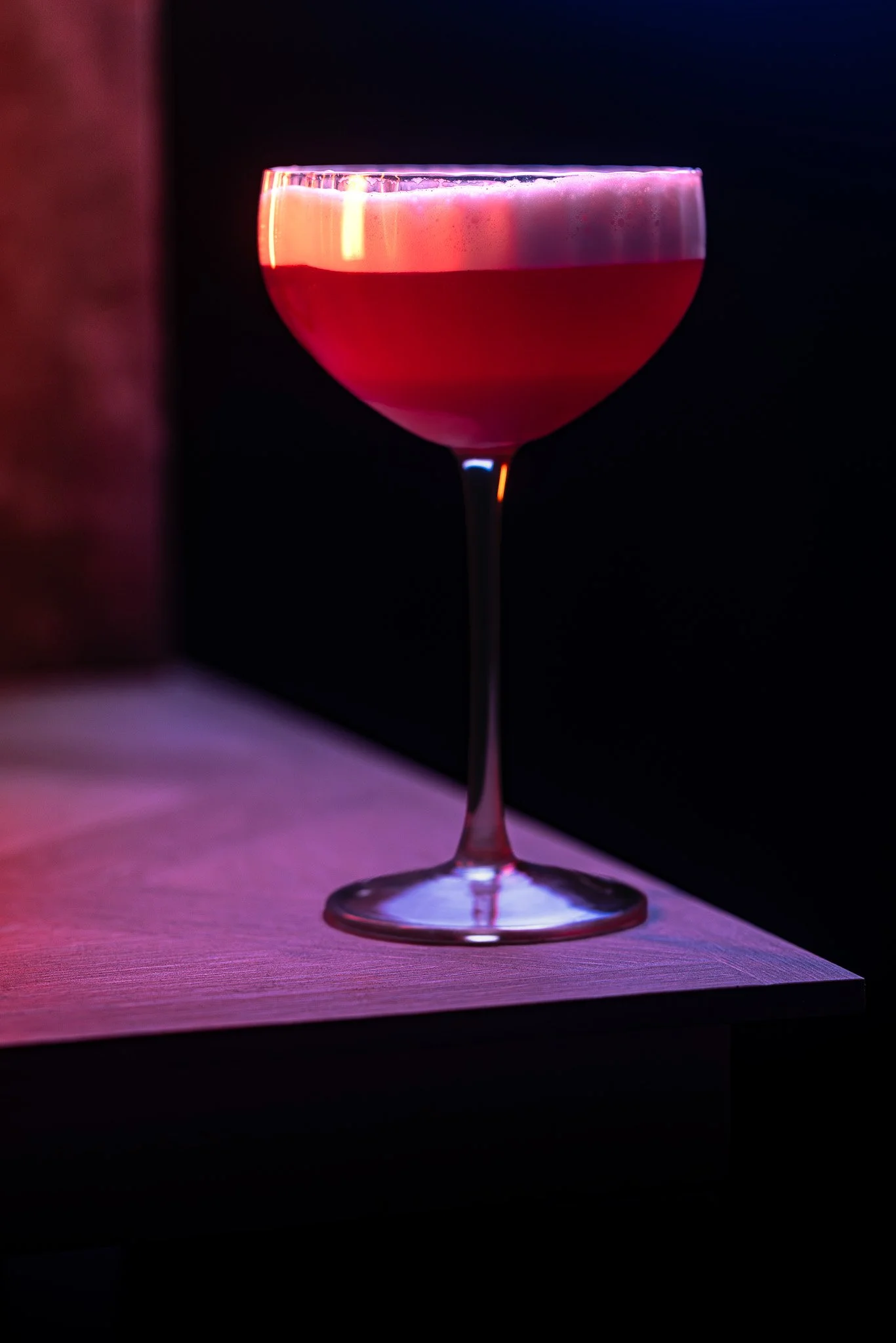 A pink cocktail in a coupe glass on a wooden surface in a dark setting.