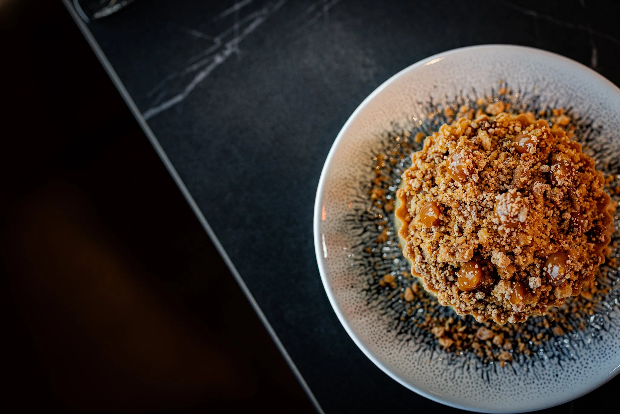 Close-up of a dessert with crumbled topping and caramel dots on a decorative ceramic plate.