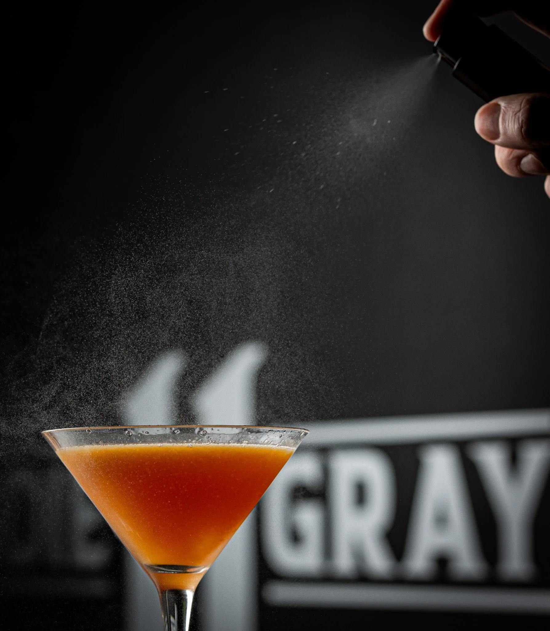 A person sprinkles a spice or sugar from a black container onto a cocktail in a martini glass, with a black and white background.