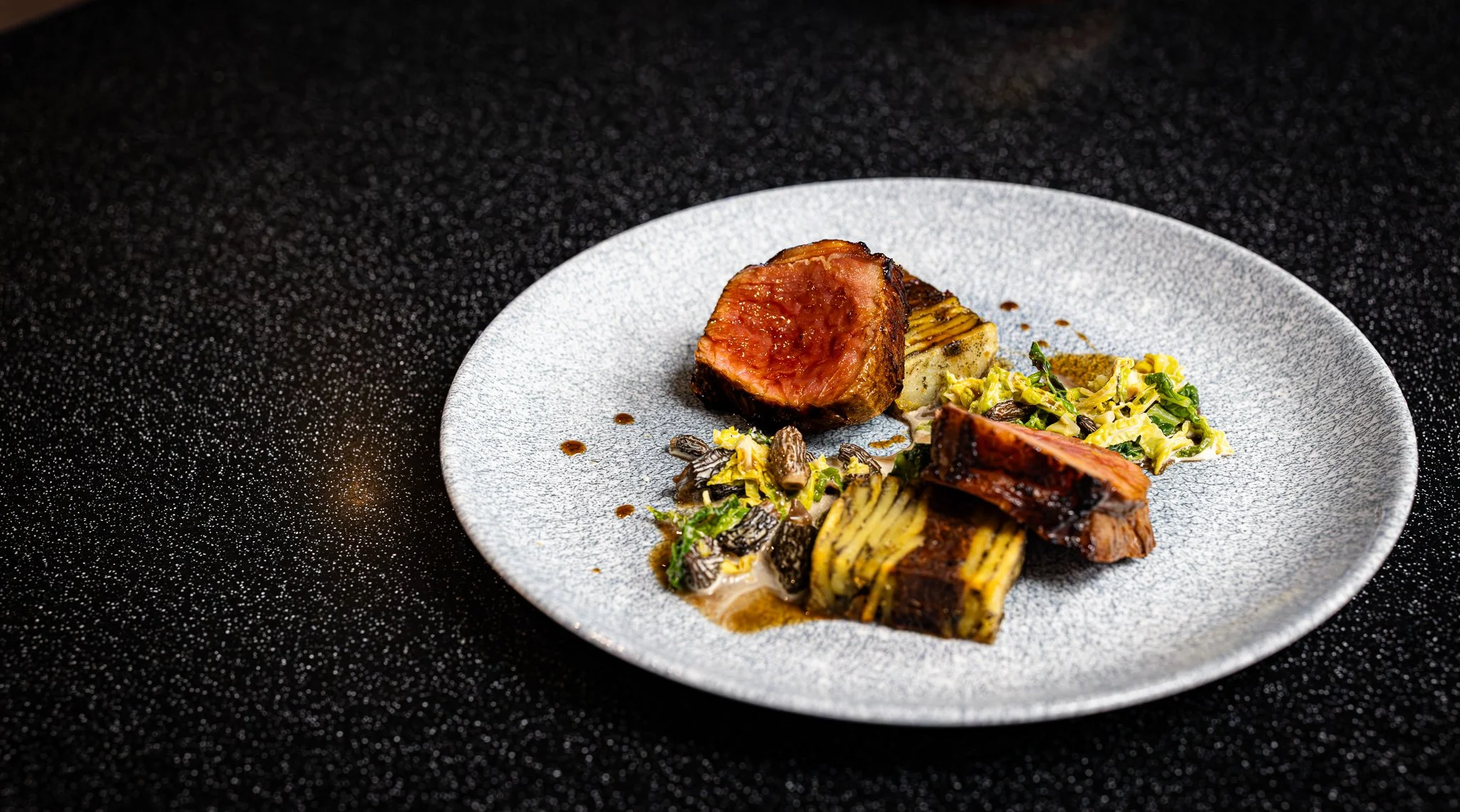 A plated gourmet meal featuring seared meat, possibly beef or lamb, garnished with vegetables and sauce, on a textured white plate against a black speckled background.