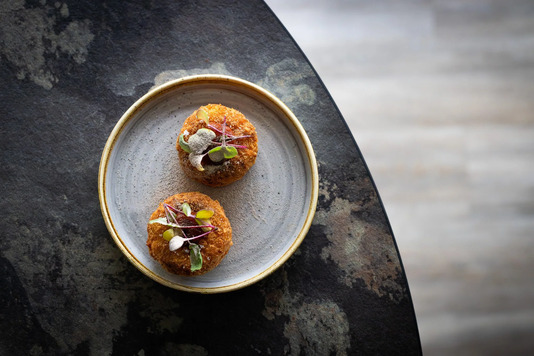 Two fried appetizers garnished with microgreens on a white plate, placed on a dark textured surface.