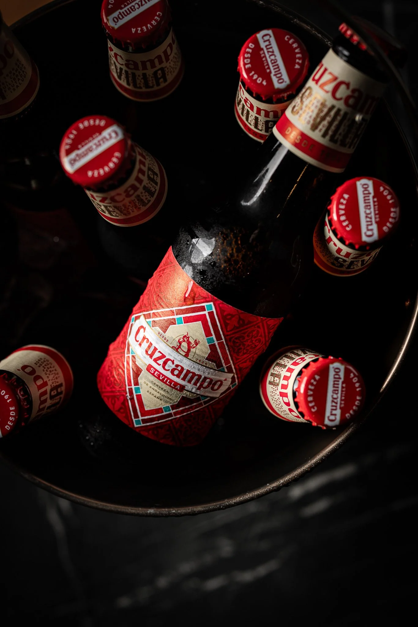 A bottle of Cruzcampo beer inside a bucket filled with ice, surrounded by several smaller cans of Cruzcampo beer.