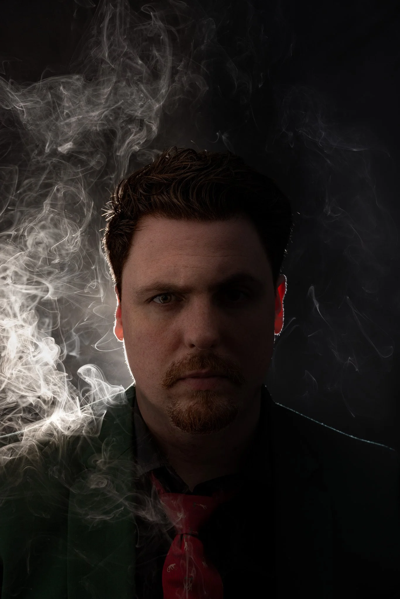 A man with a mustache and goatee, wearing a dark suit and red tie, is in front of a dark background with swirling smoke around his head.