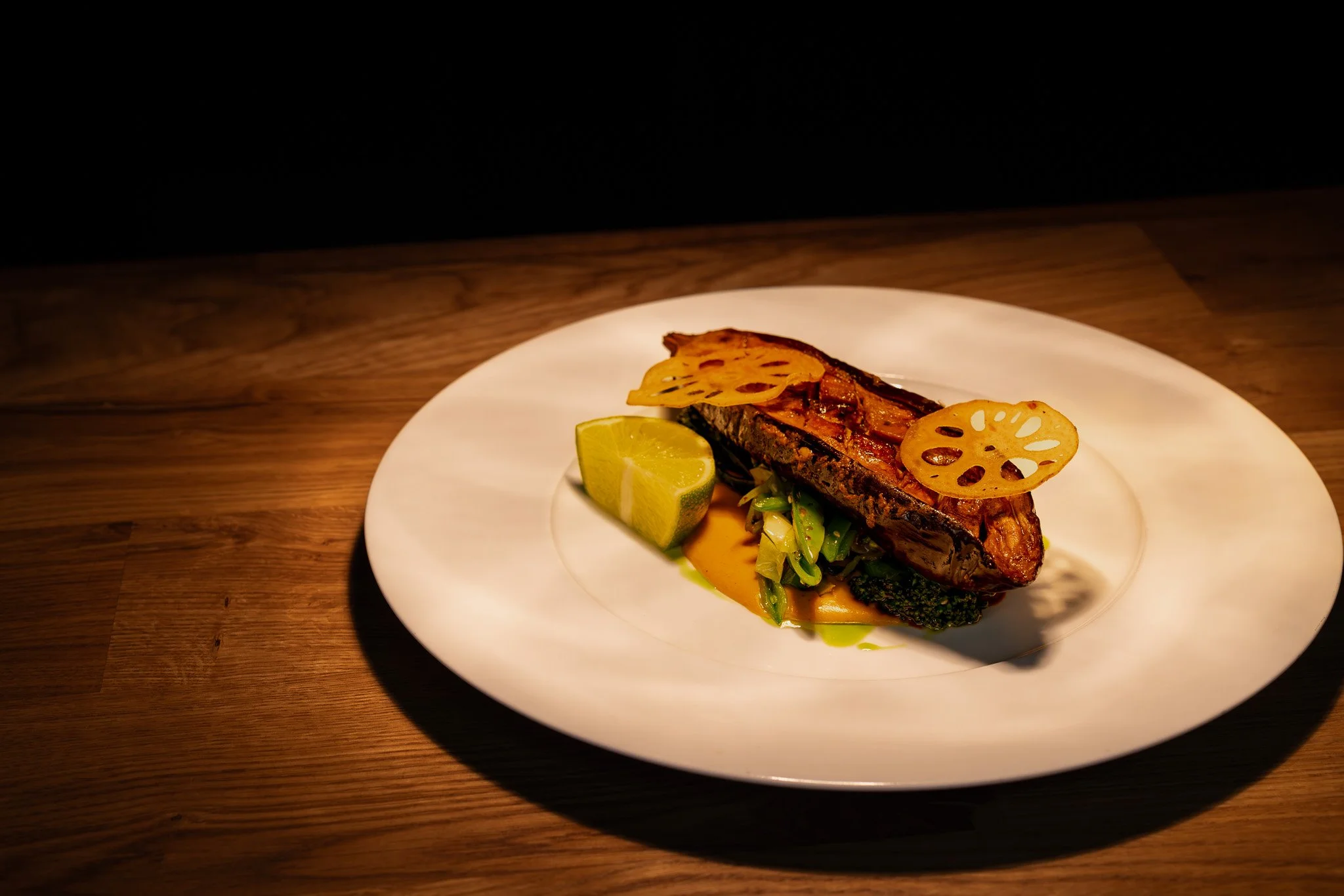Grilled fish fillet with lotus root chips, lemon wedge, greens, and sauce on a white plate.
